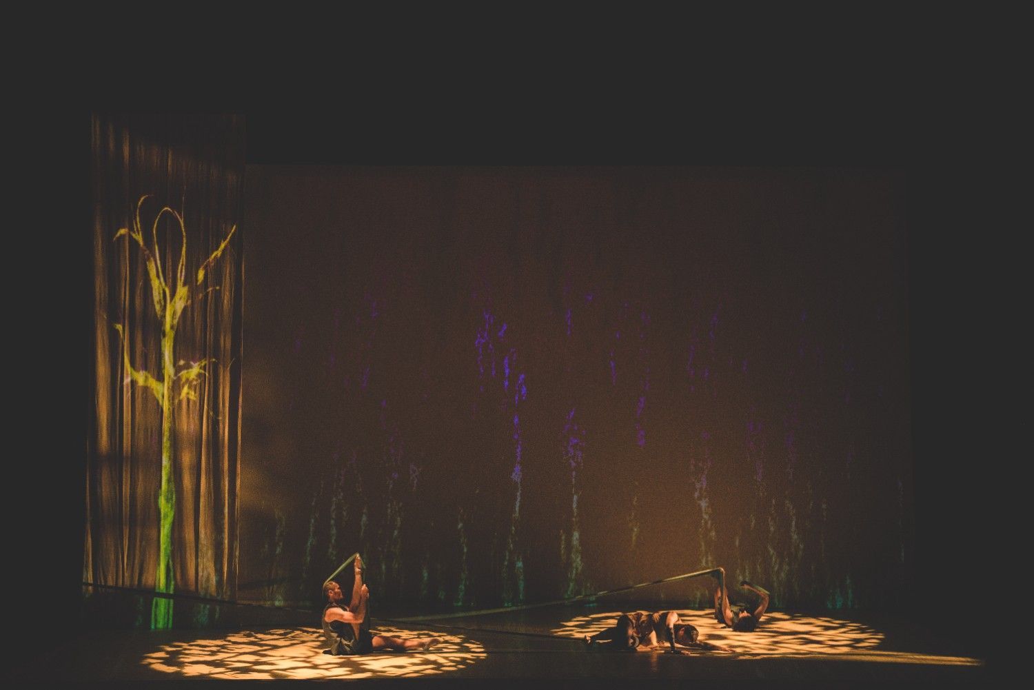 Escenario con poca luz, dos artistas sentados bajo focos cálidos y un elemento escénico alto y brillante a la izquierda.
