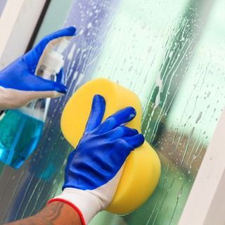 Manos con guantes azules limpiando una superficie de vidrio mojada con una esponja amarilla y un pulverizador