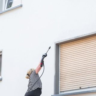 Una persona limpiando con una hidrolavadora la pared blanca de un edificio, junto a una ventana grande con la persiana bajada