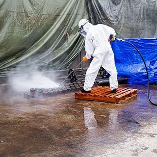 Un trabajador con un traje protector blanco lava a presión una superficie mojada junto a una lona azul.