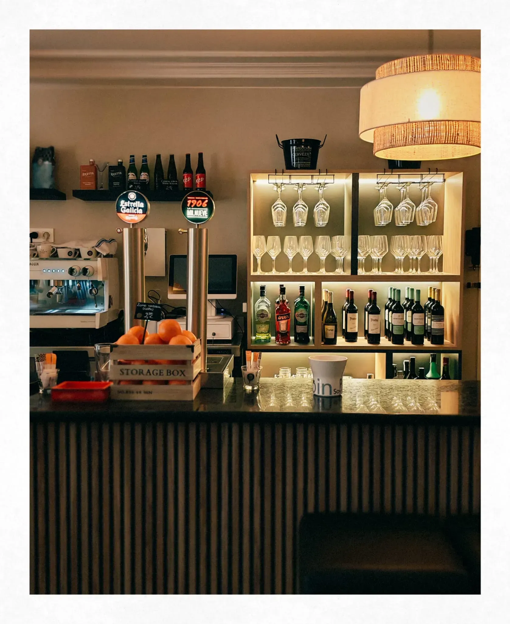 Acogedora barra de bar con botellas, copas colgantes, naranjas y una cálida iluminación colgante