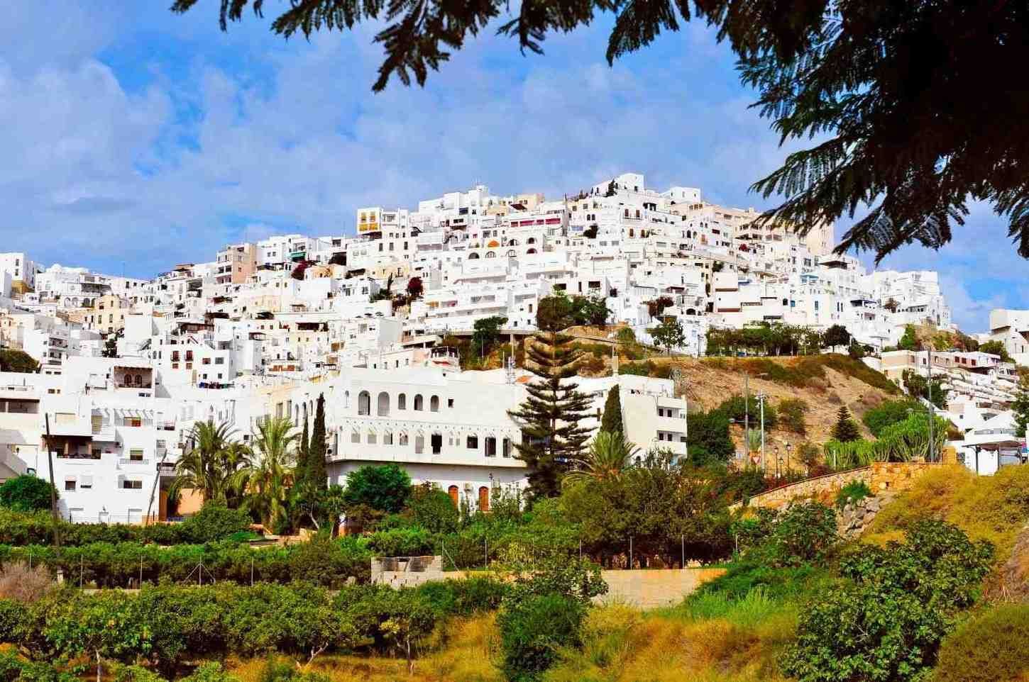 Pueblo blanco en la ladera de una colina con casas coloridas, terrazas verdes y cielo azul enmarcado por ramas de árboles.