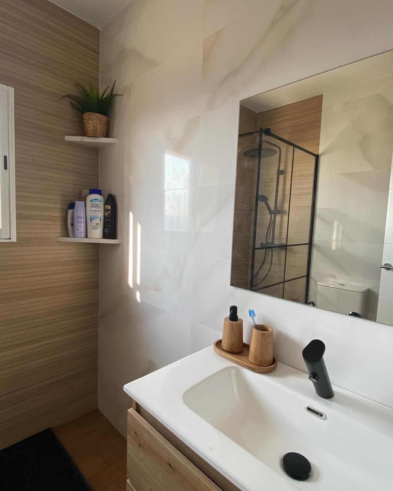 Lavabo moderno con espejo, paredes beige, reflejo de la ducha y una pequeña repisa con artículos de tocador y plantas.
