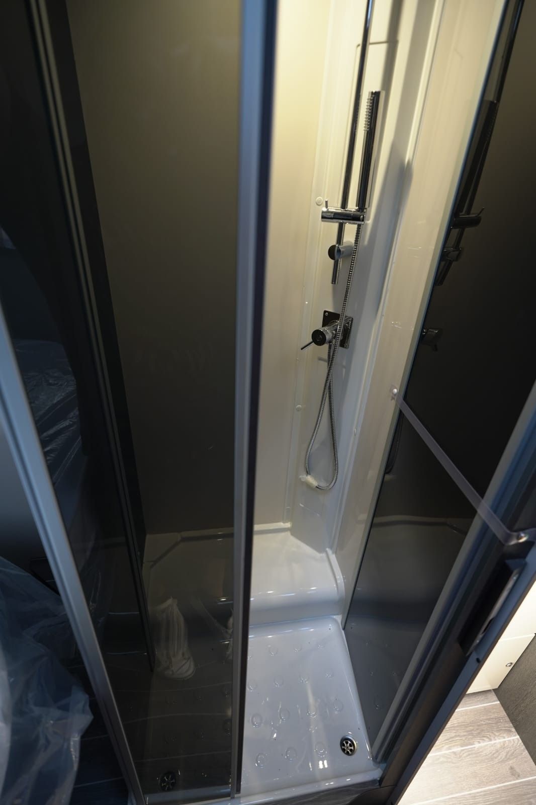 Pequeña cabina de ducha acristalada con suelo de baldosas blancas y una manguera de ducha en la pared.