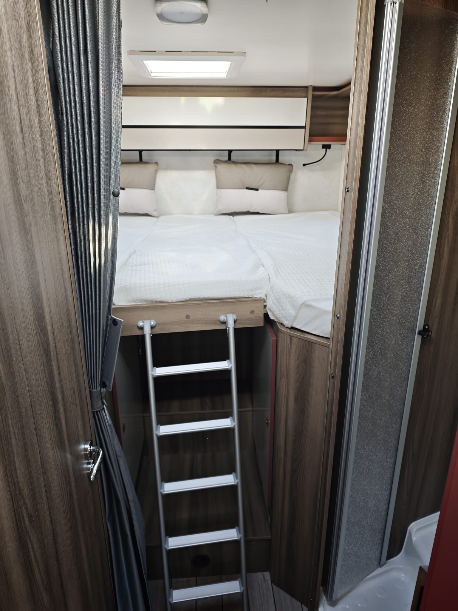Dormitorio estrecho de caravana con cama, escalera y armarios beige a lo largo de las paredes.