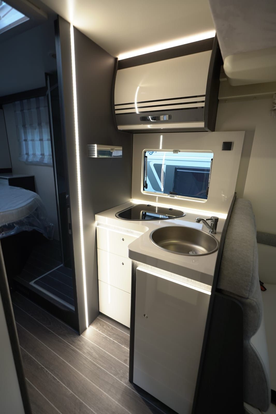 Cocina compacta para autocaravana con fregadero, microondas, armario y suelo de madera oscura en un interior estrecho.