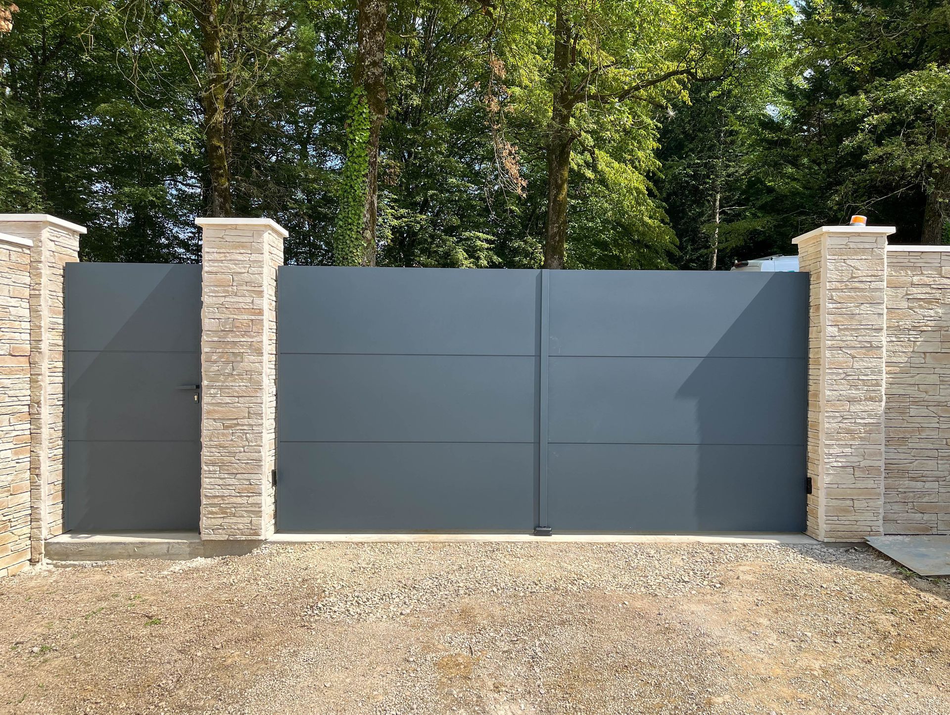 Portail moderne en panneaux gris pleins, encadré par deux piliers en pierre faisant partie d’un mur en pierre. Sol en gravier à l’avant et végétation en arrière-plan.