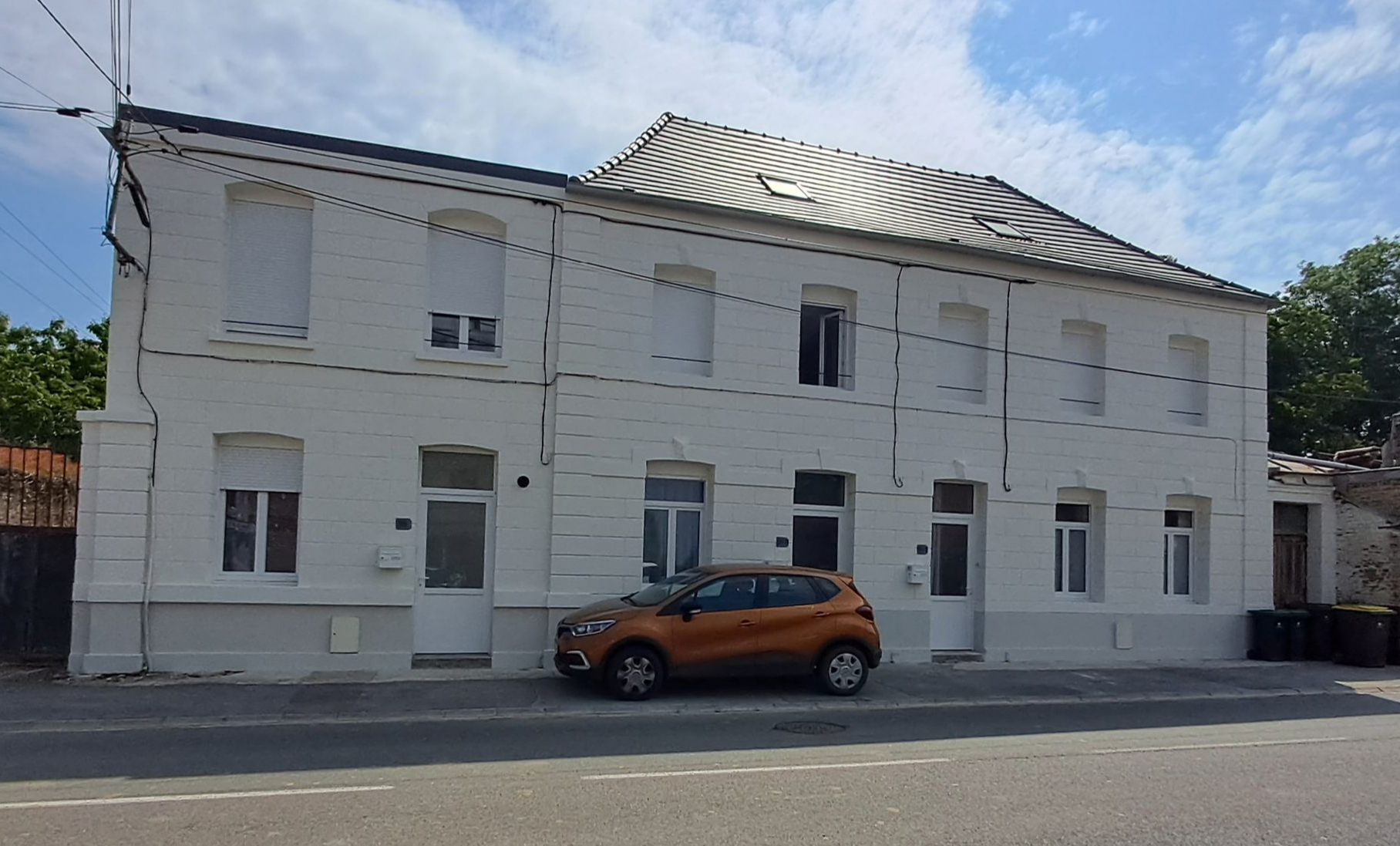 Bâtiment blanc de deux étages avec de nombreuses fenêtres et portes ; une voiture orange garée devant.