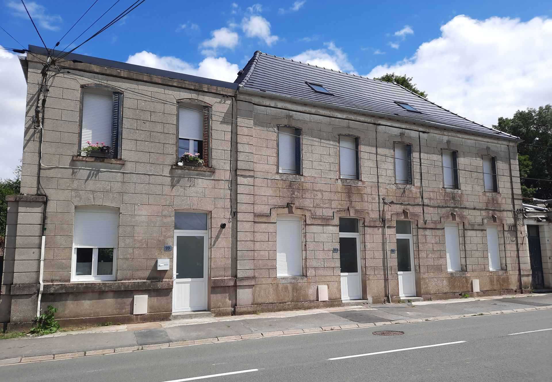 Bâtiment en pierre de deux étages avec plusieurs portes et fenêtres blanches ; ciel bleu au-dessus.