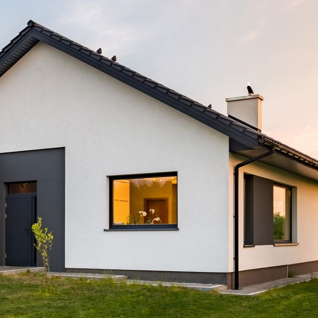 Maison blanche moderne avec boiseries, encadrement de fenêtre et de porte foncés ; jardin gazonné ; coucher de soleil.