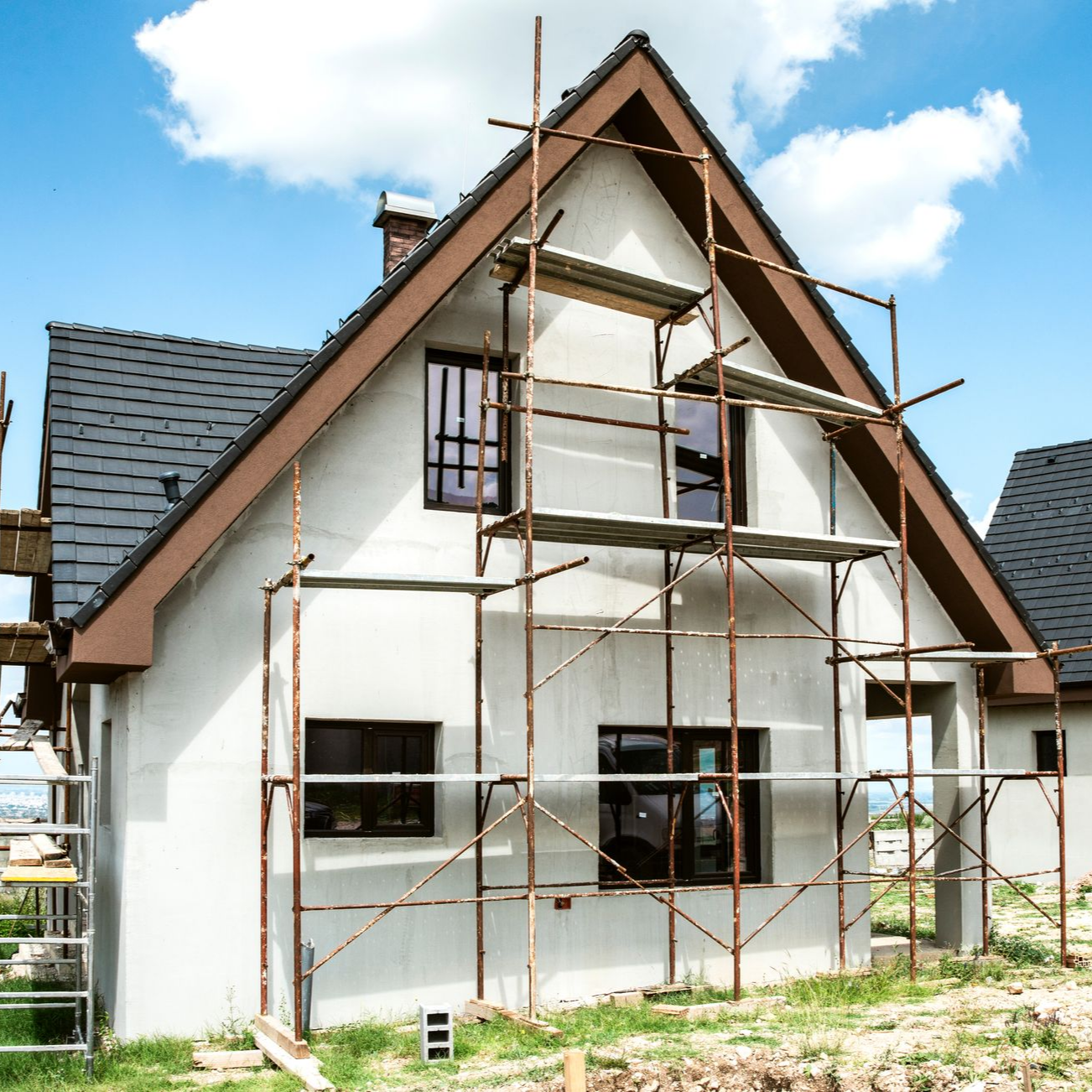 Maison en construction avec échafaudages devant une façade en stuc blanc. Boiseries marron, toit noir.