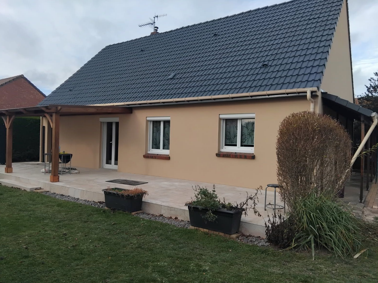 Maison beige avec toit, fenêtres et terrasse gris. Pelouse verte au premier plan.