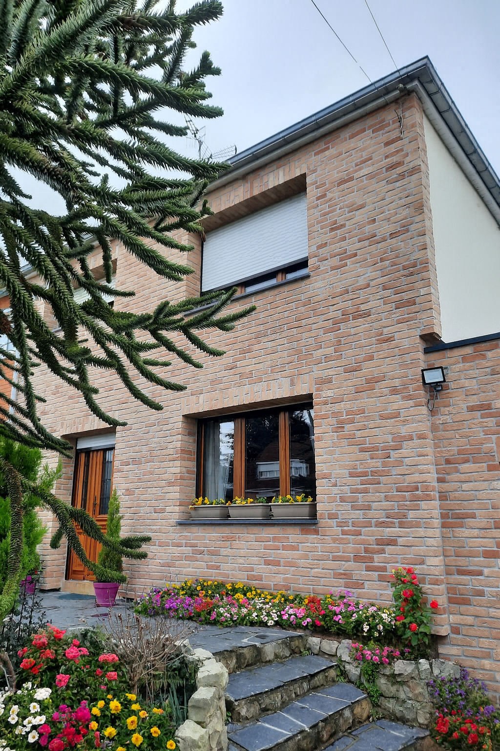 Maison en briques avec escalier, un parterre de fleurs colorées et des fenêtres à stores blancs.