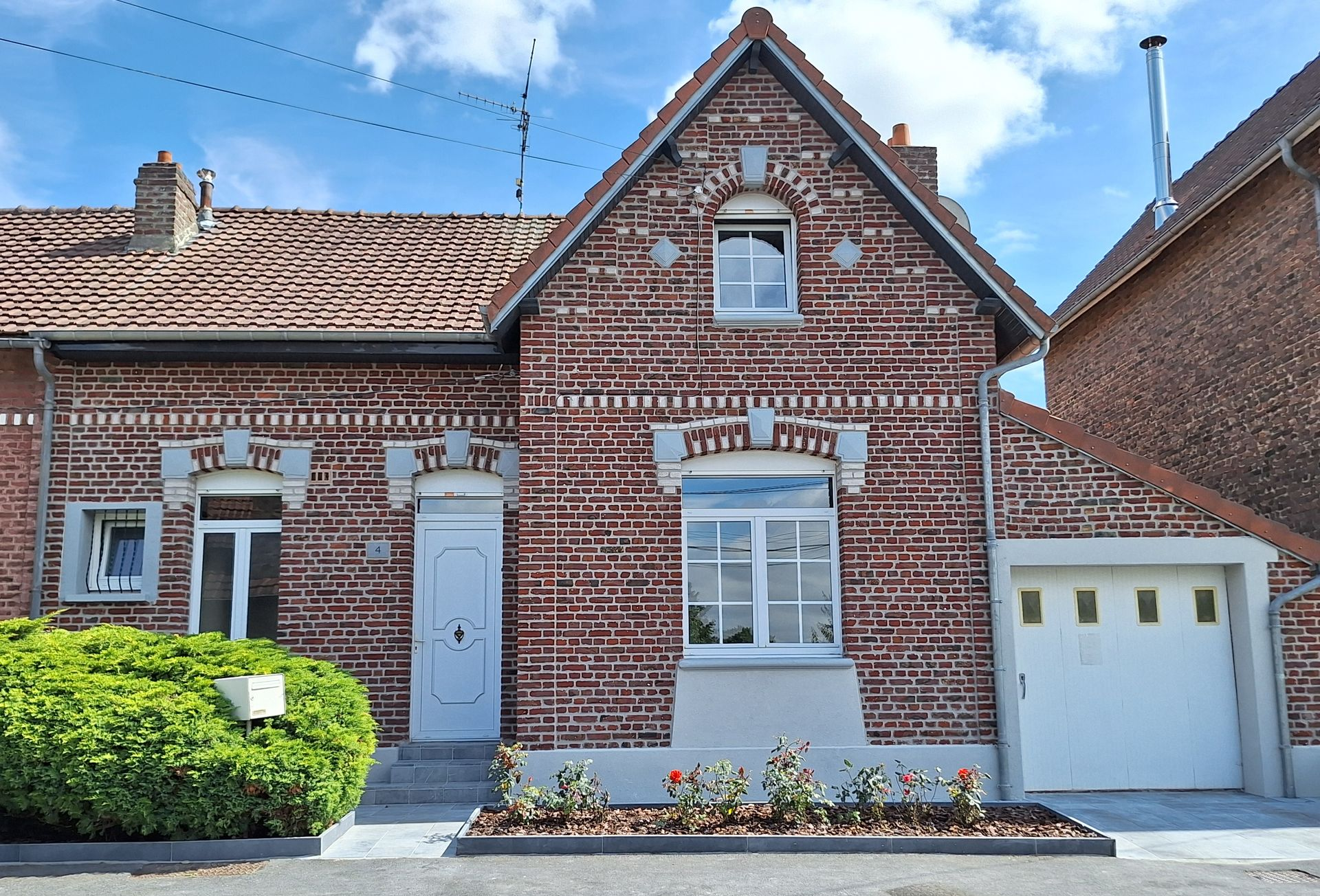 Maison en briques avec toit à pignons, boiseries blanches, petit jardin et garage attenant, sous un ciel bleu.