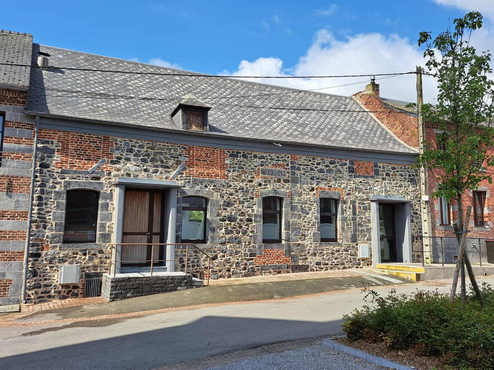 Bâtiment en pierre et en brique, toit en ardoise, trois fenêtres et porte ouverte, situé dans une rue en légère pente.