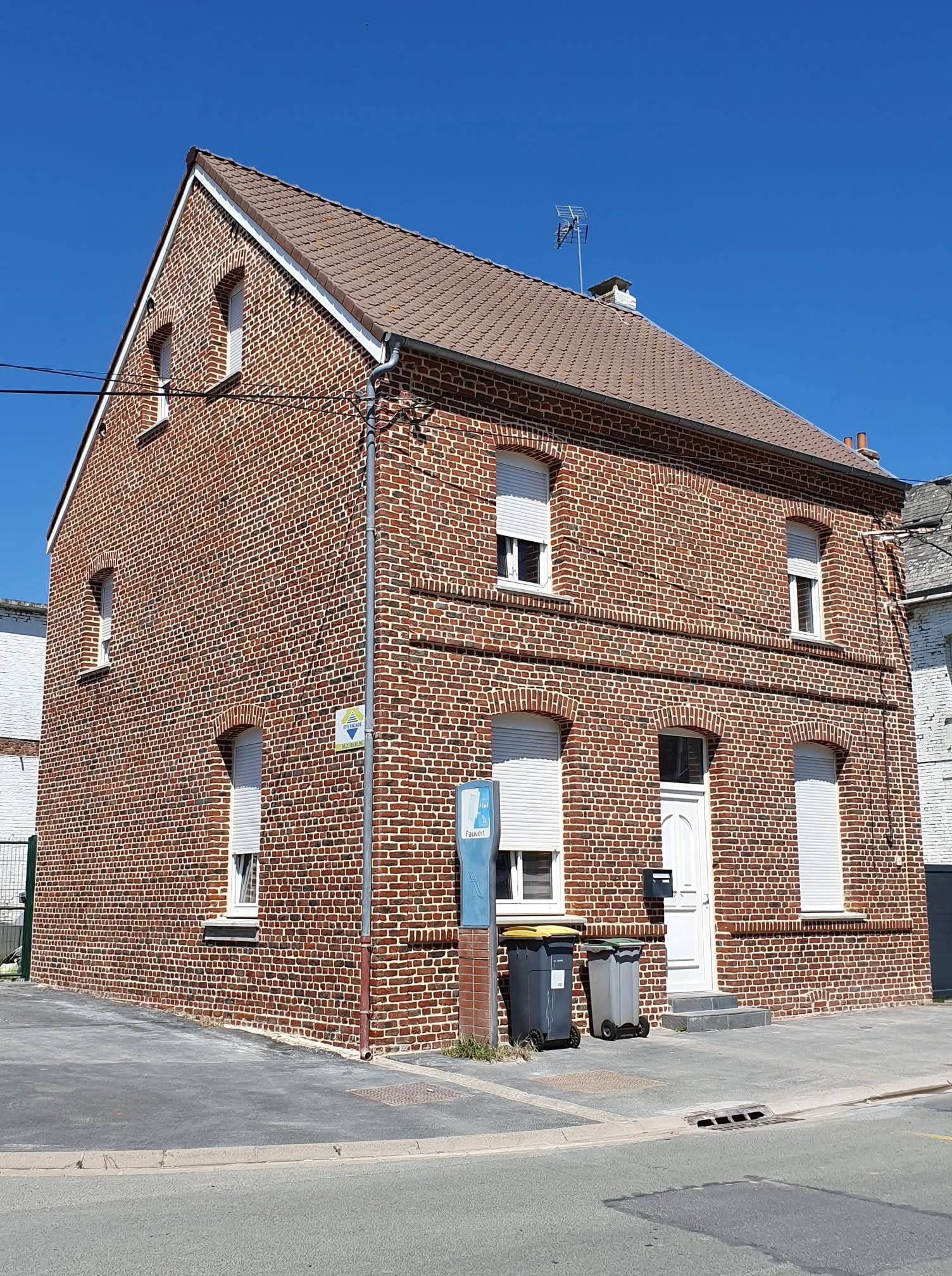 Maison en briques de deux étages avec un toit à deux pans, de nombreuses fenêtres à volets blancs, une porte blanche et des poubelles.