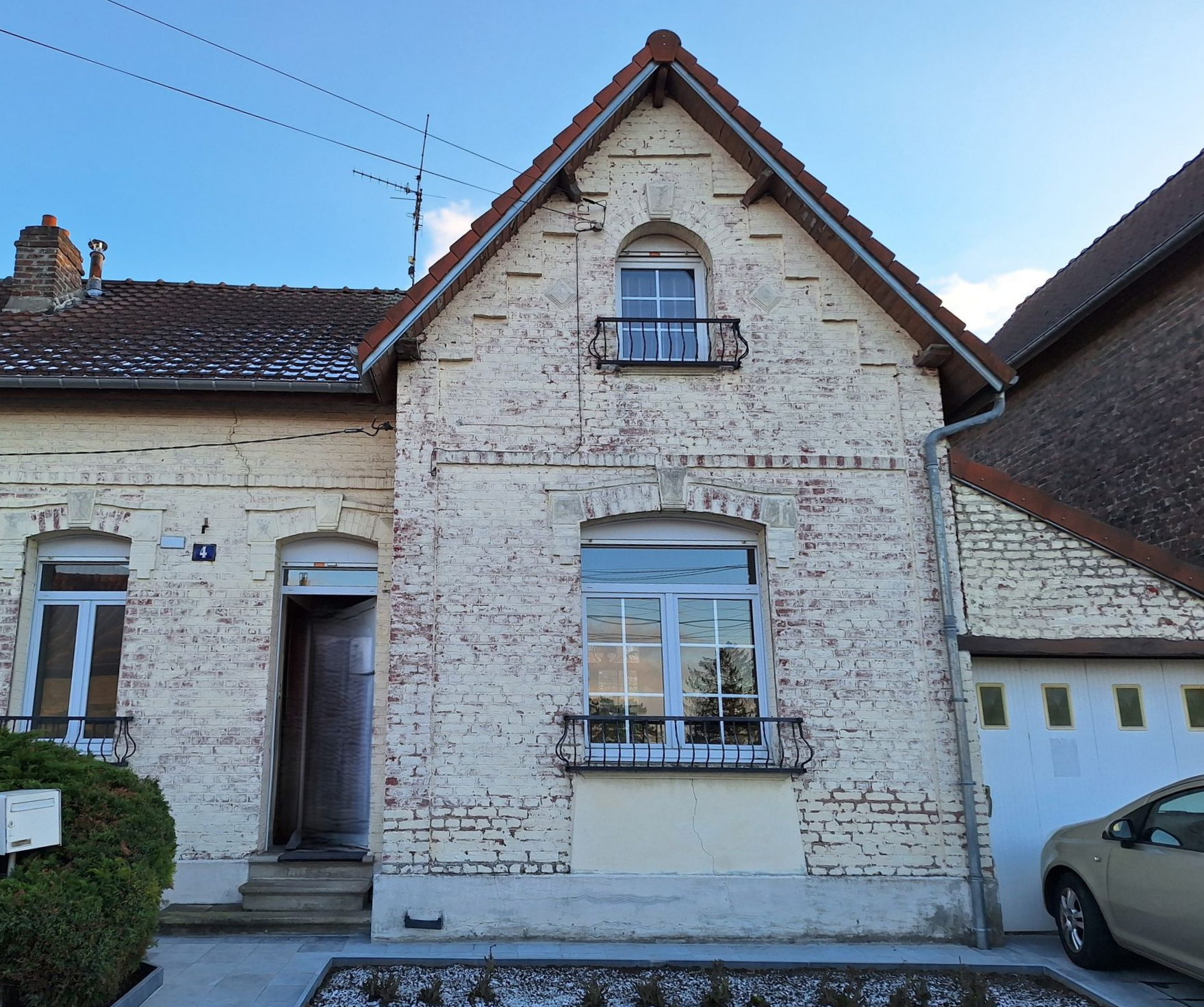 Maison en briques avec pignon arqué, deux fenêtres avec petits balcons, voiture garée à côté.
