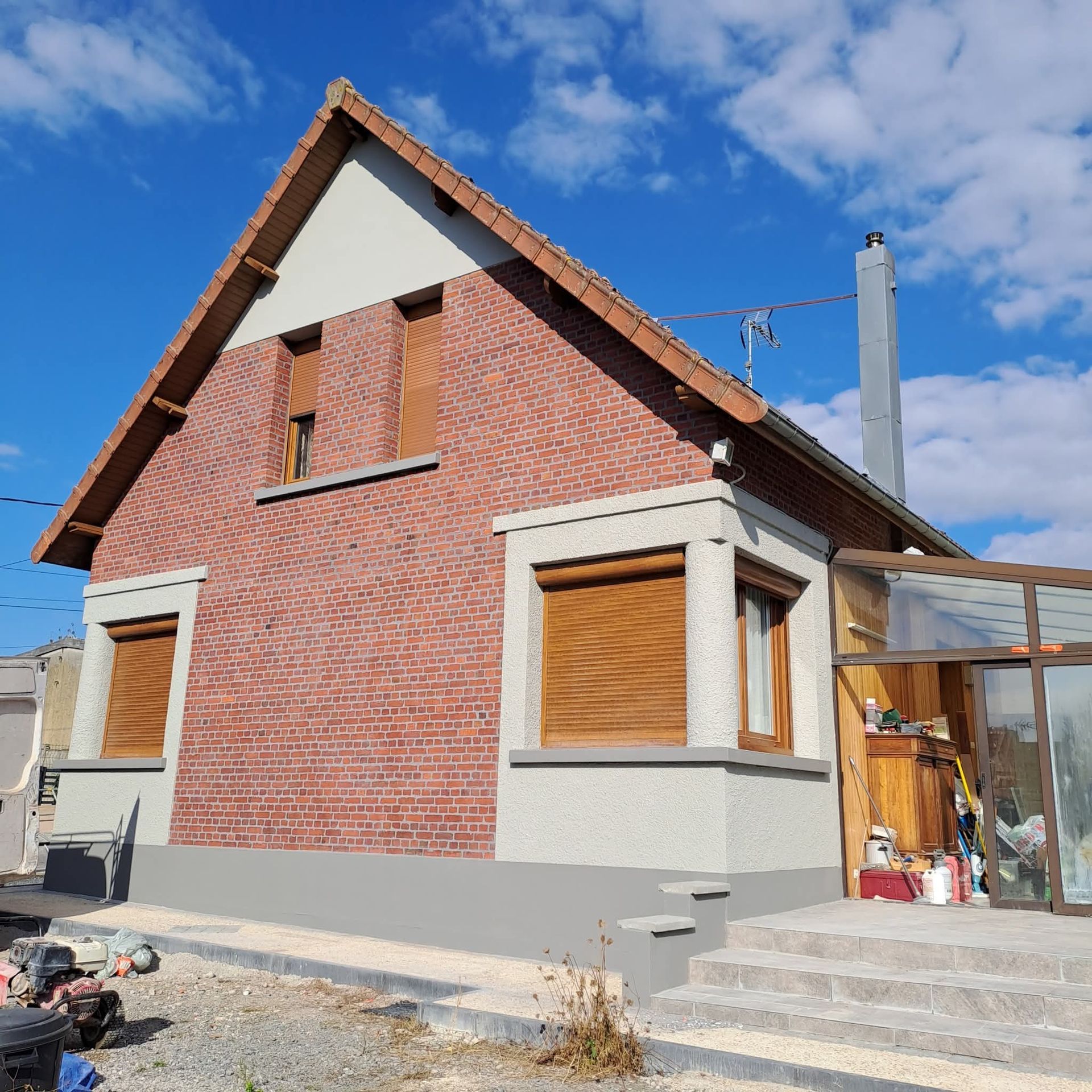 Maison en briques avec volets en bois, cheminée et véranda vitrée, se détachant sur un ciel bleu.