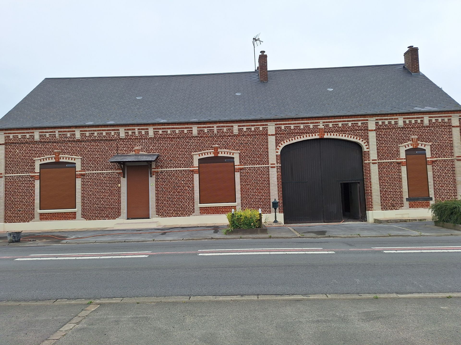 Bâtiment en briques avec un toit sombre, une grande porte noire, des volets et une route devant.