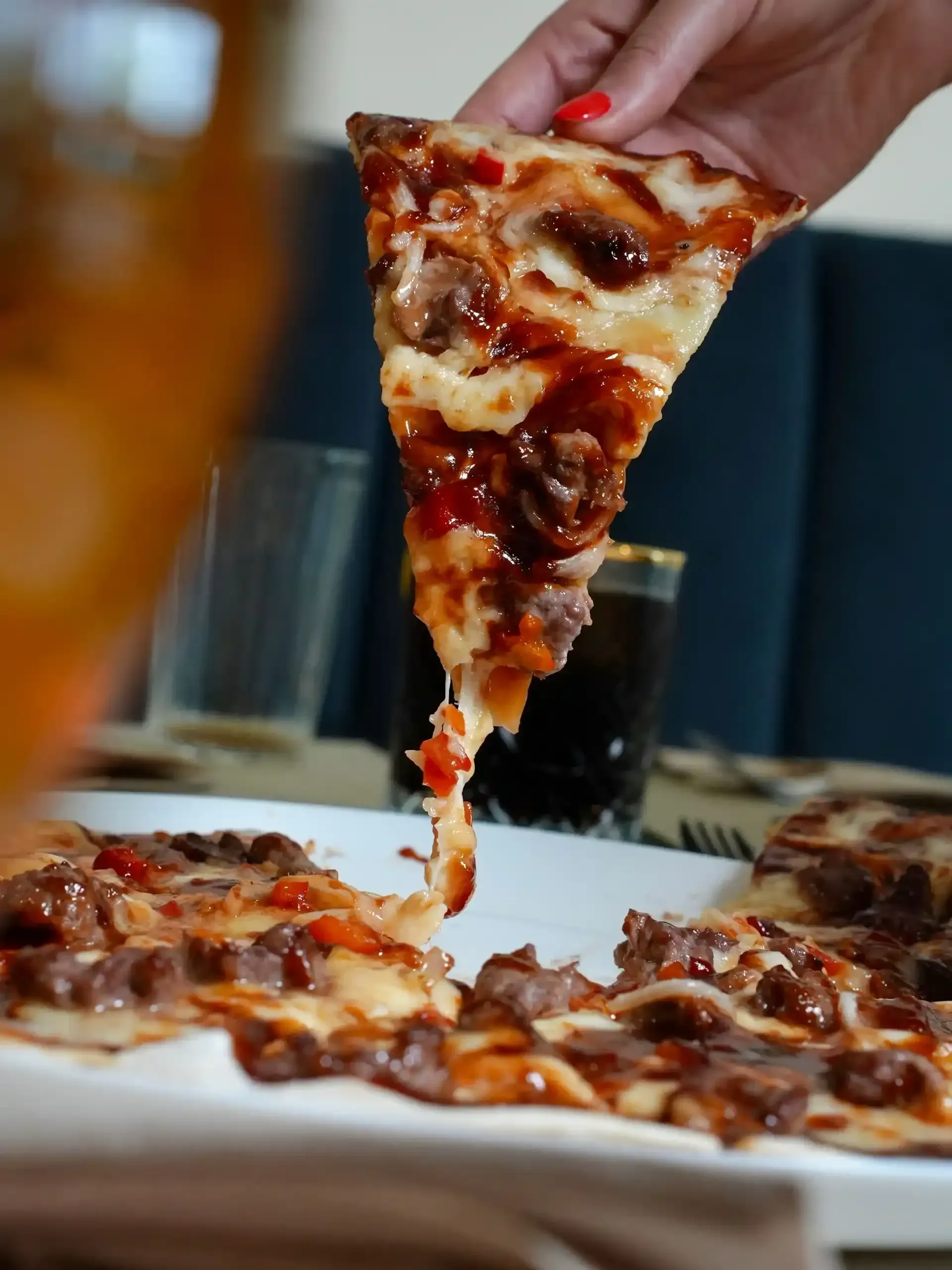 Una mano sostiene una rebanada de pizza con queso derretido, ingredientes y salsa. La pizza reposa en un plato blanco.