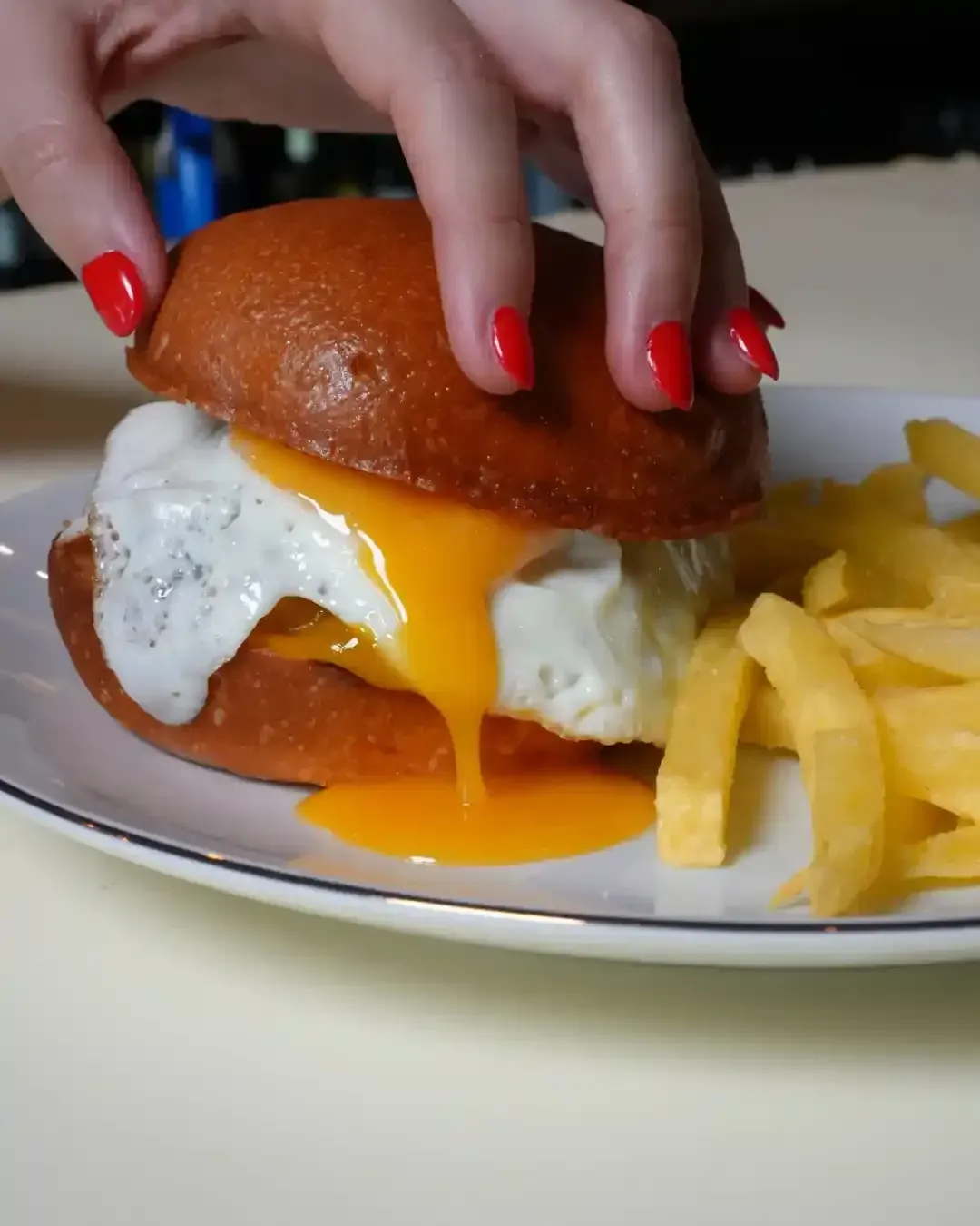 Mano de una persona con uñas rojas sosteniendo un sándwich de huevo. La yema se derrama sobre el plato con papas fritas.