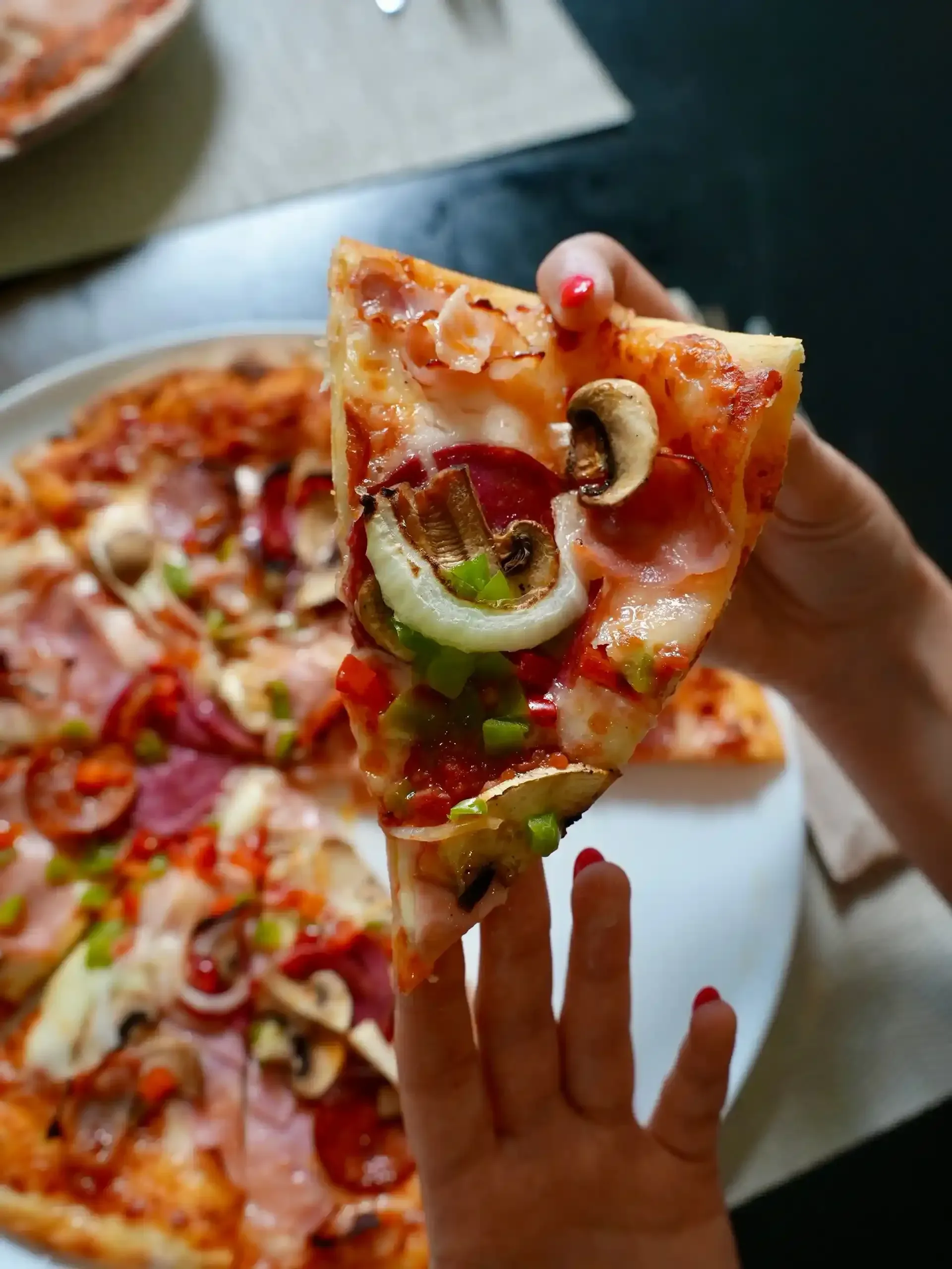 Persona sosteniendo una porción de pizza con ingredientes visibles: champiñones, cebollas, pepperoni y pimientos verdes.