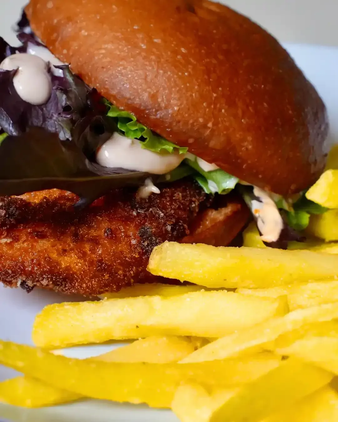 Sándwich de pollo crujiente con lechuga, salsa y papas fritas en un plato blanco.