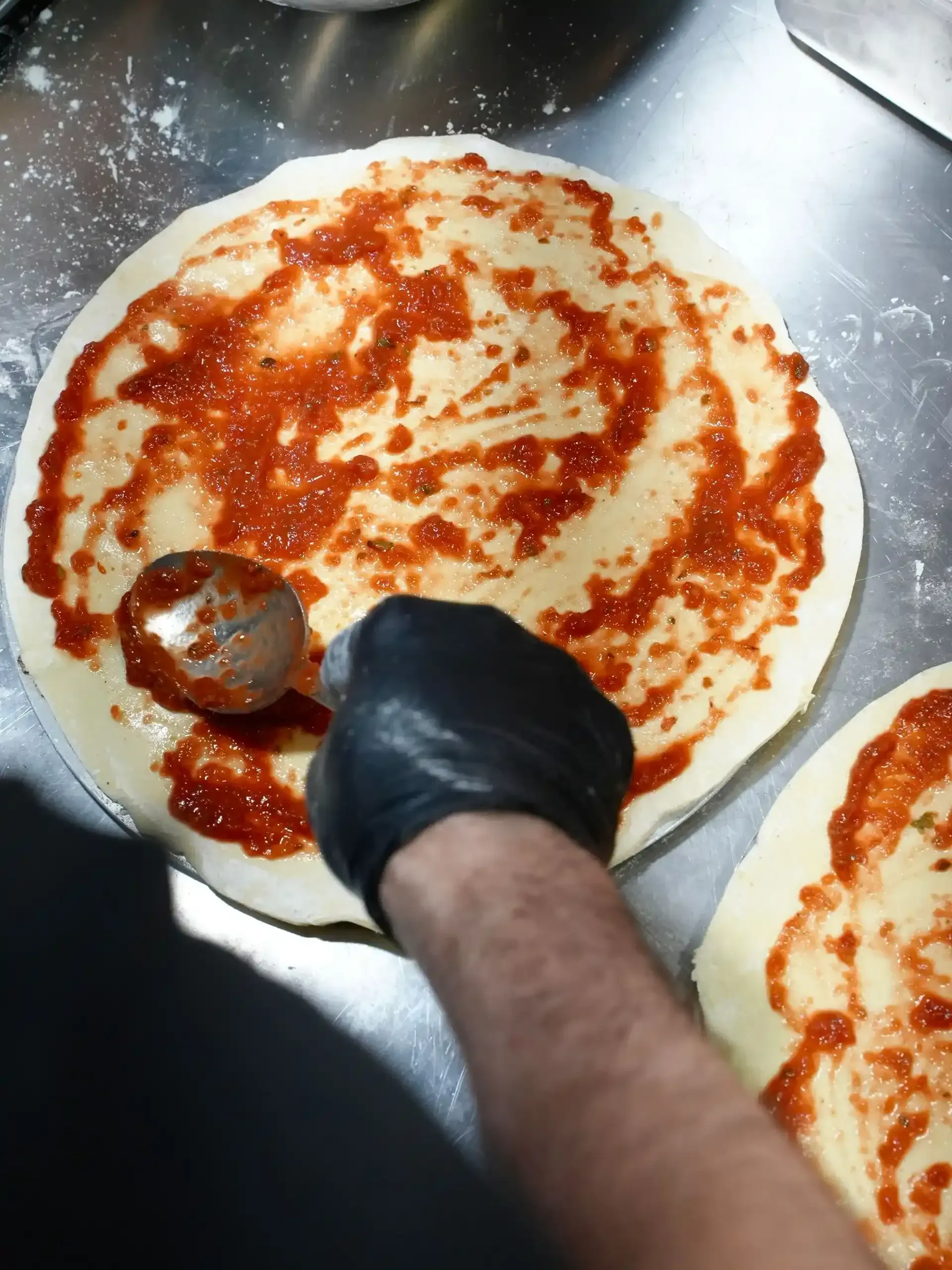 Persona untando salsa de tomate sobre masa de pizza con una cuchara. Mano enguantada negra.