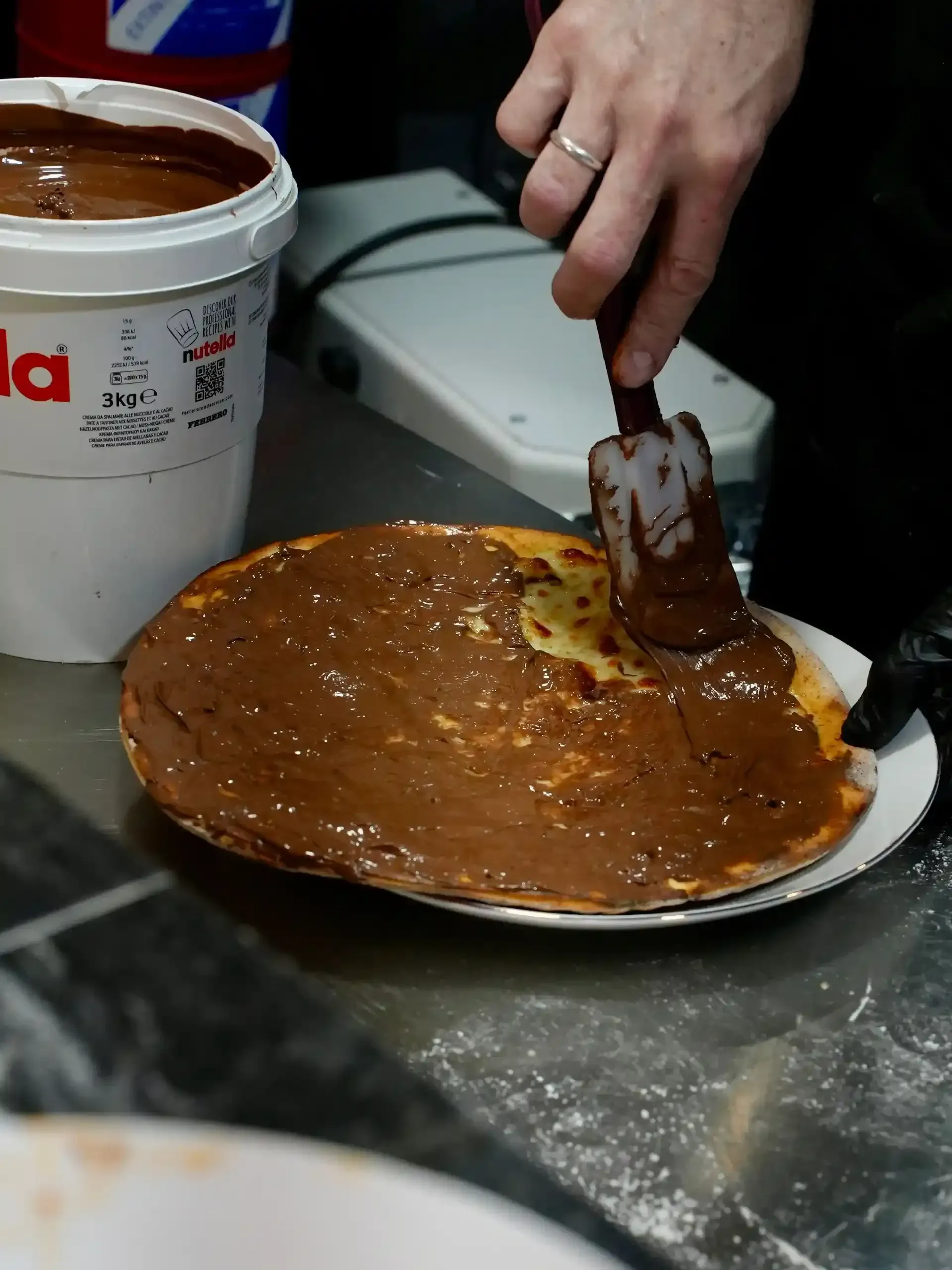 Una persona extiende crema de chocolate y avellanas con una espátula sobre un panqueque grande.