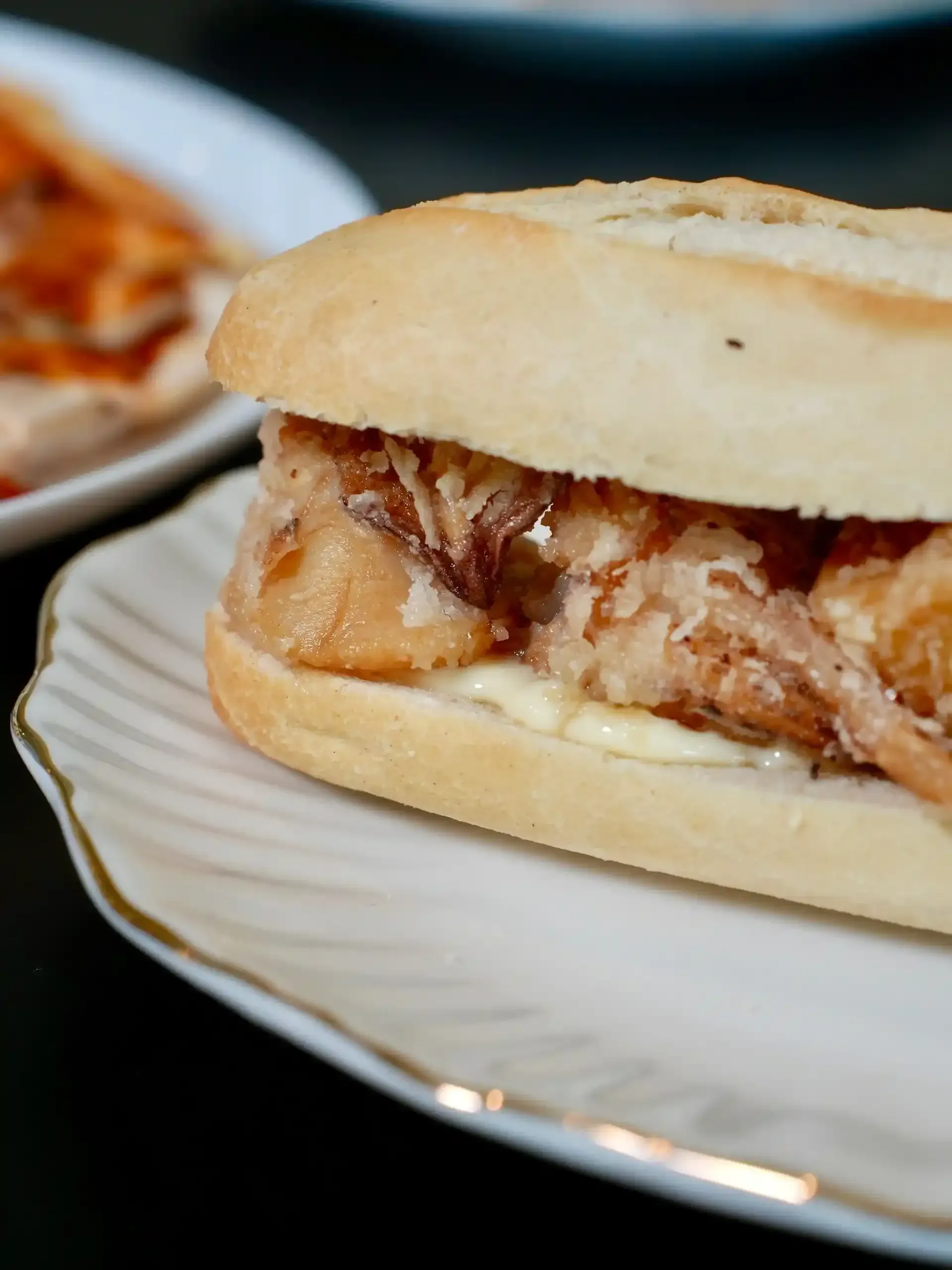 Sándwich en plato blanco con borde dorado; pan de color claro, relleno de comida frita y mayonesa.