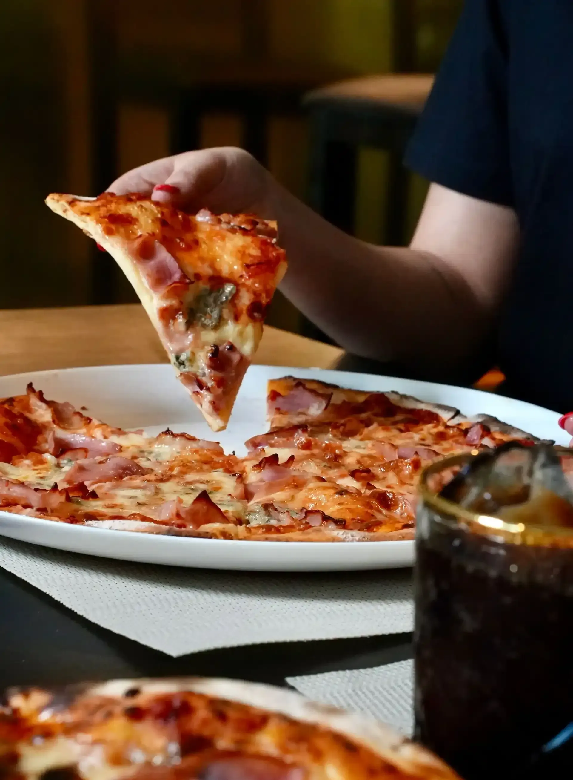 Persona sosteniendo una porción de pizza sobre un plato de pizza, con un refresco cerca.