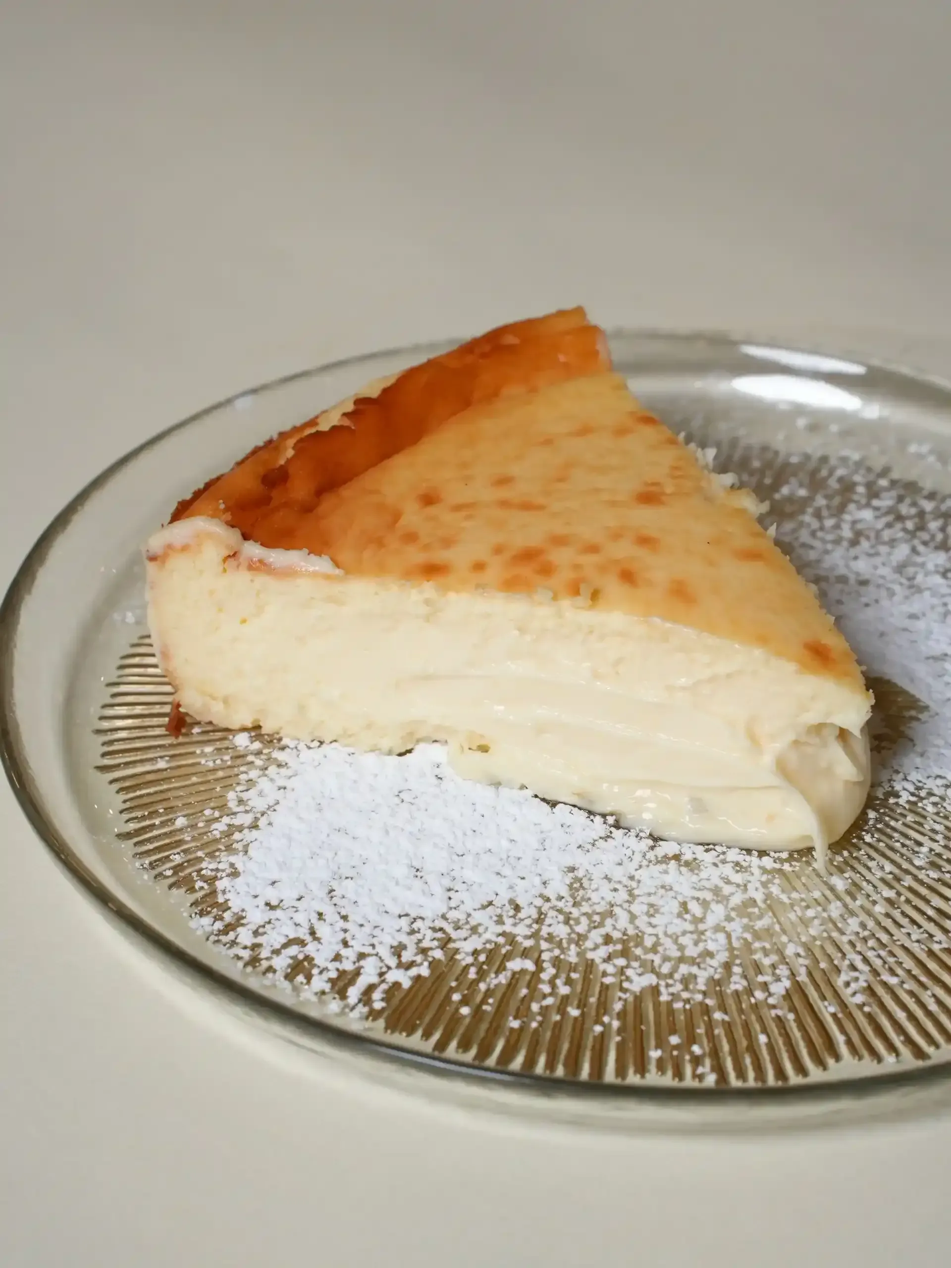 Rebanada de tarta de queso en un plato espolvoreada con azúcar glas, bizcocho de color crema claro y parte superior de color marrón dorado.