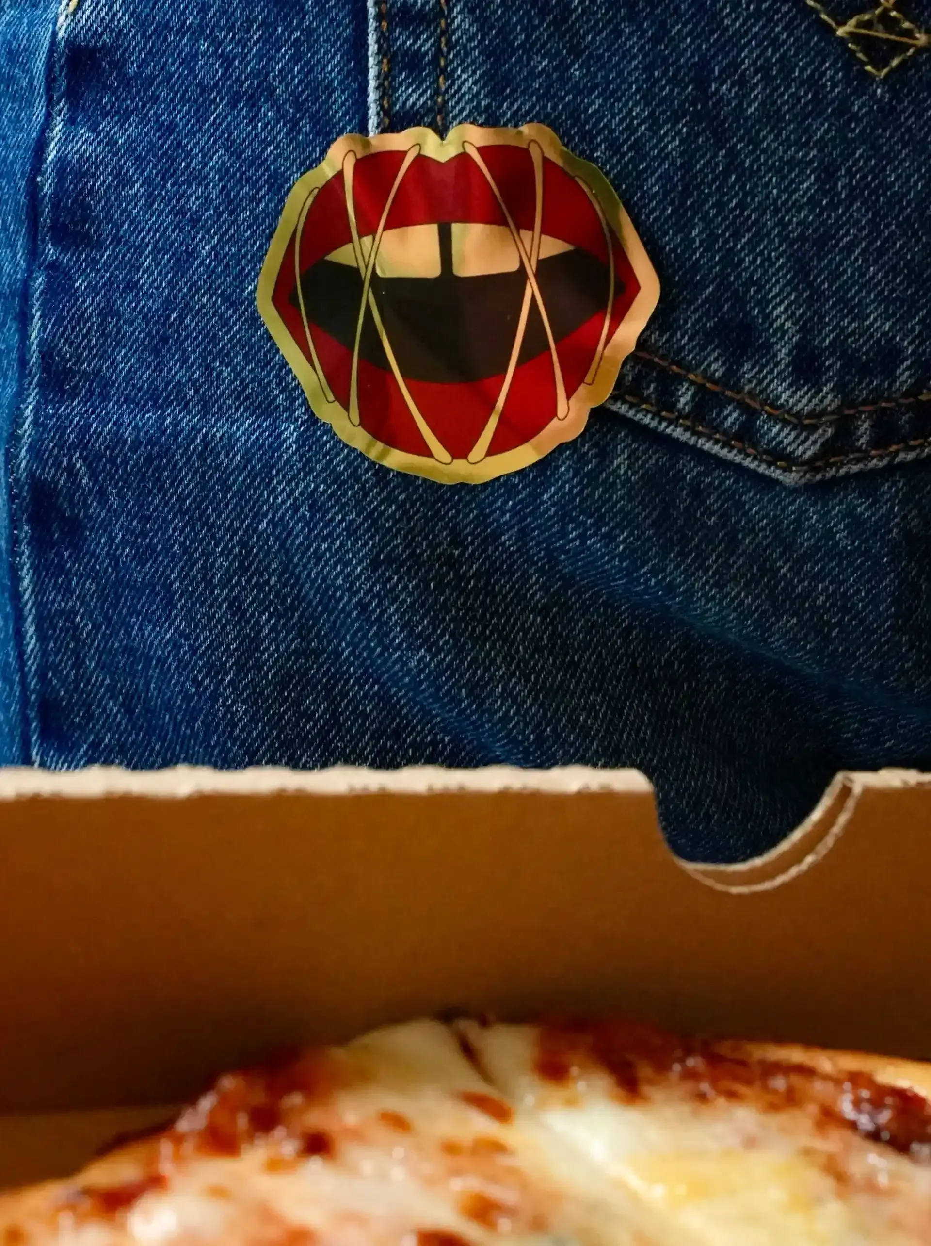 Pin con diseño de boca sonriente en un bolsillo de mezclilla azul con una caja de pizza visible debajo.
