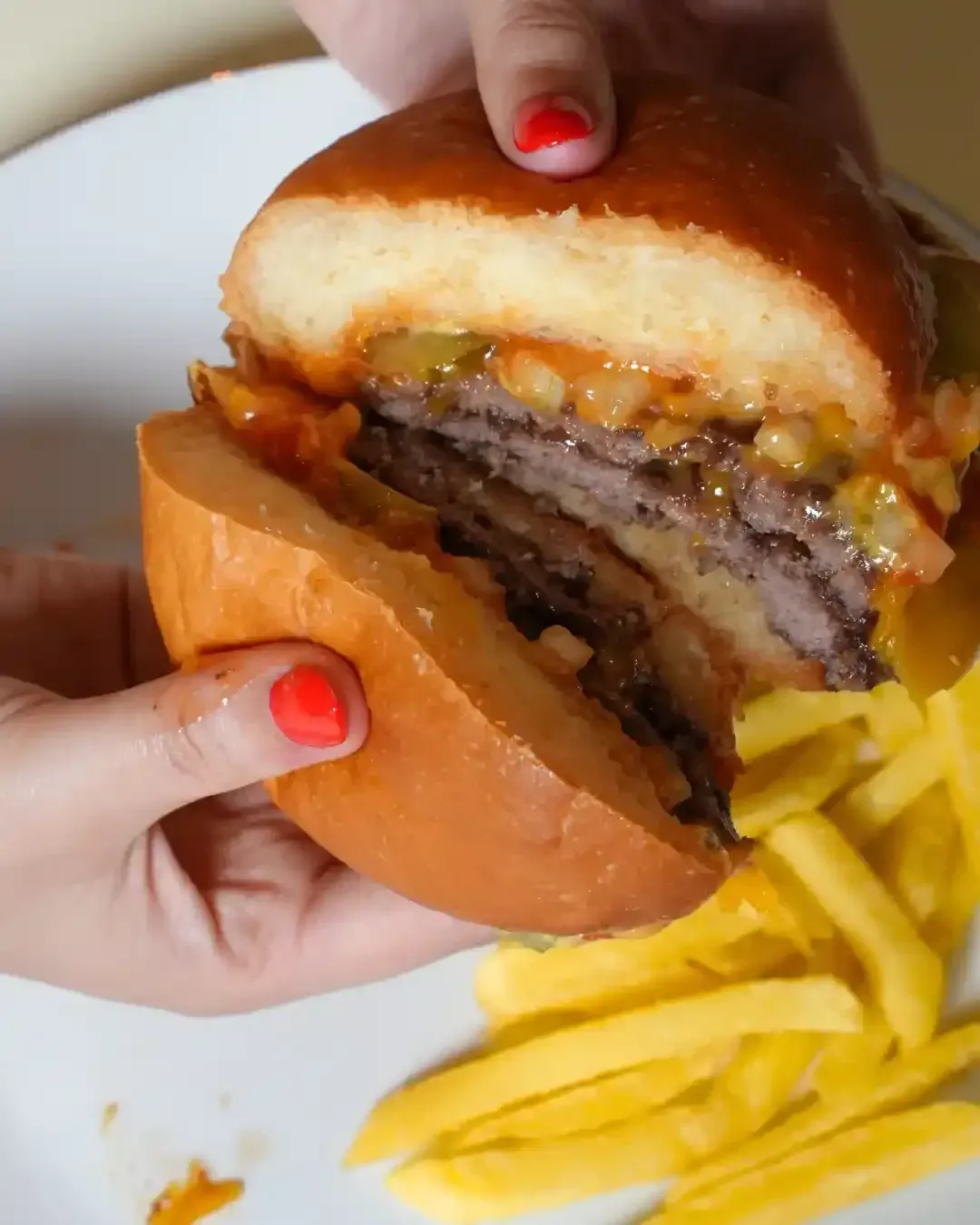Persona sosteniendo una hamburguesa cortada por la mitad, revelando dos piezas con sus ingredientes. Se ven papas fritas en el plato.