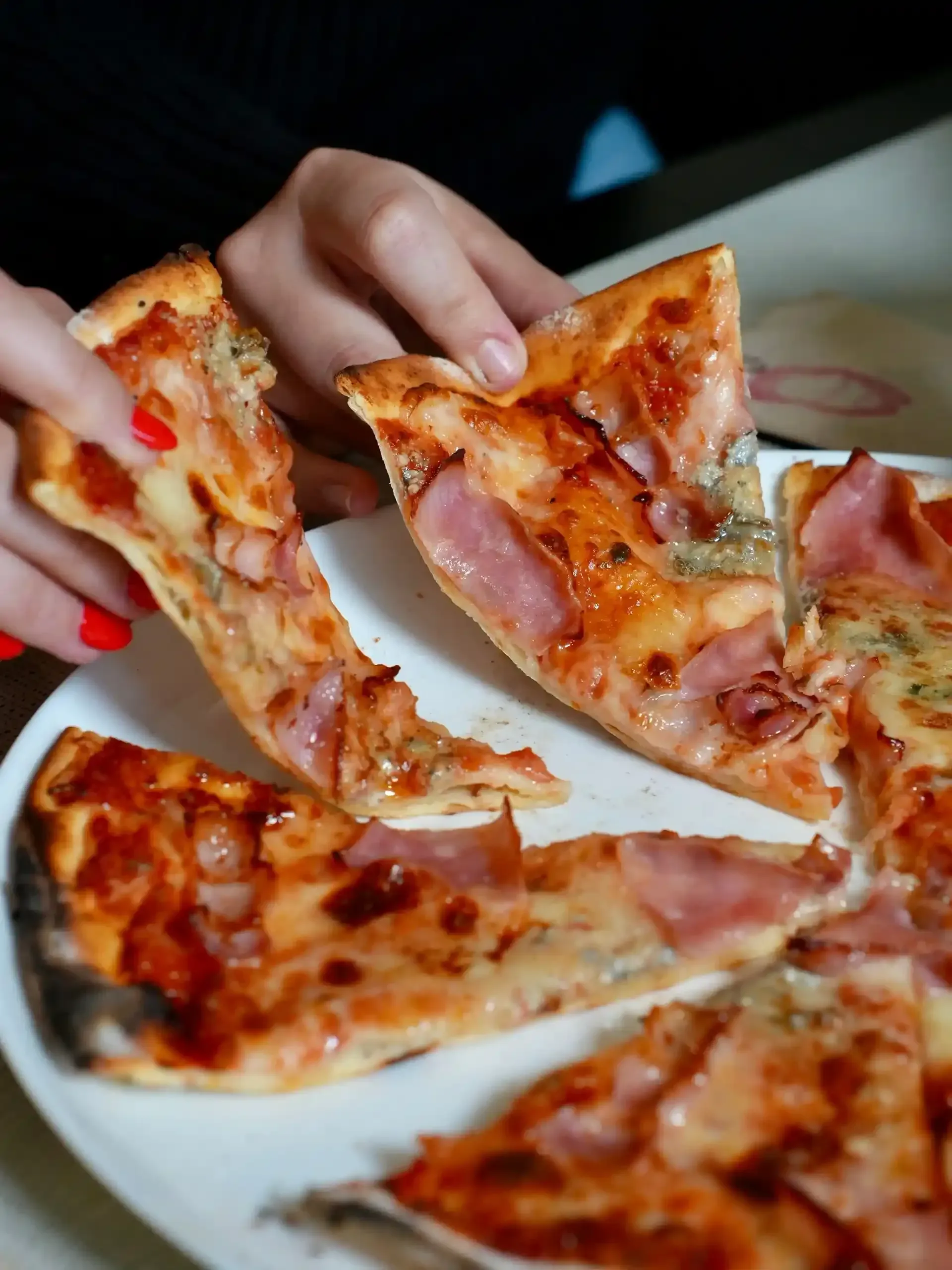 Manos de una persona sosteniendo porciones de pizza con jamón y queso en un plato blanco.