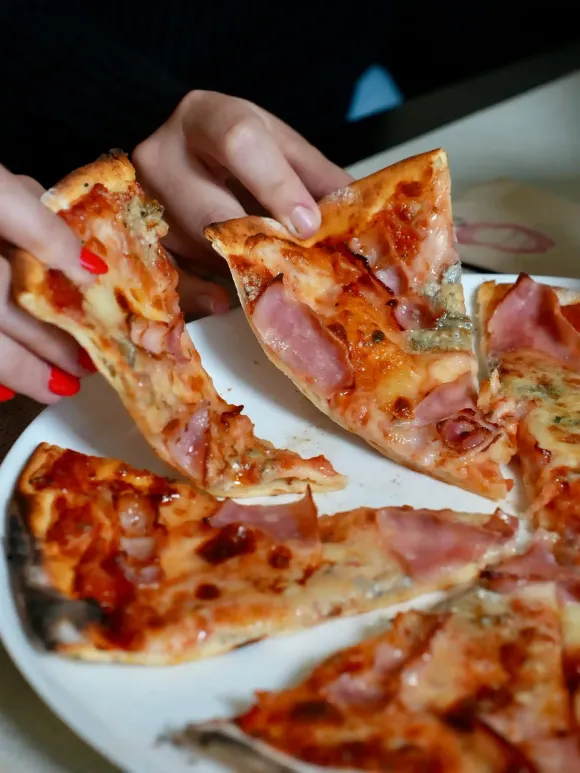 Manos de una persona sosteniendo porciones de pizza con jamón y queso en un plato blanco.