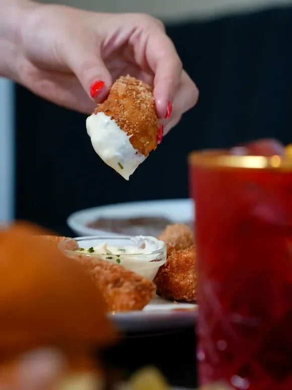 Persona mojando un alimento empanizado en un tazón pequeño de salsa blanca. Cerca hay una bebida roja y otros alimentos similares.