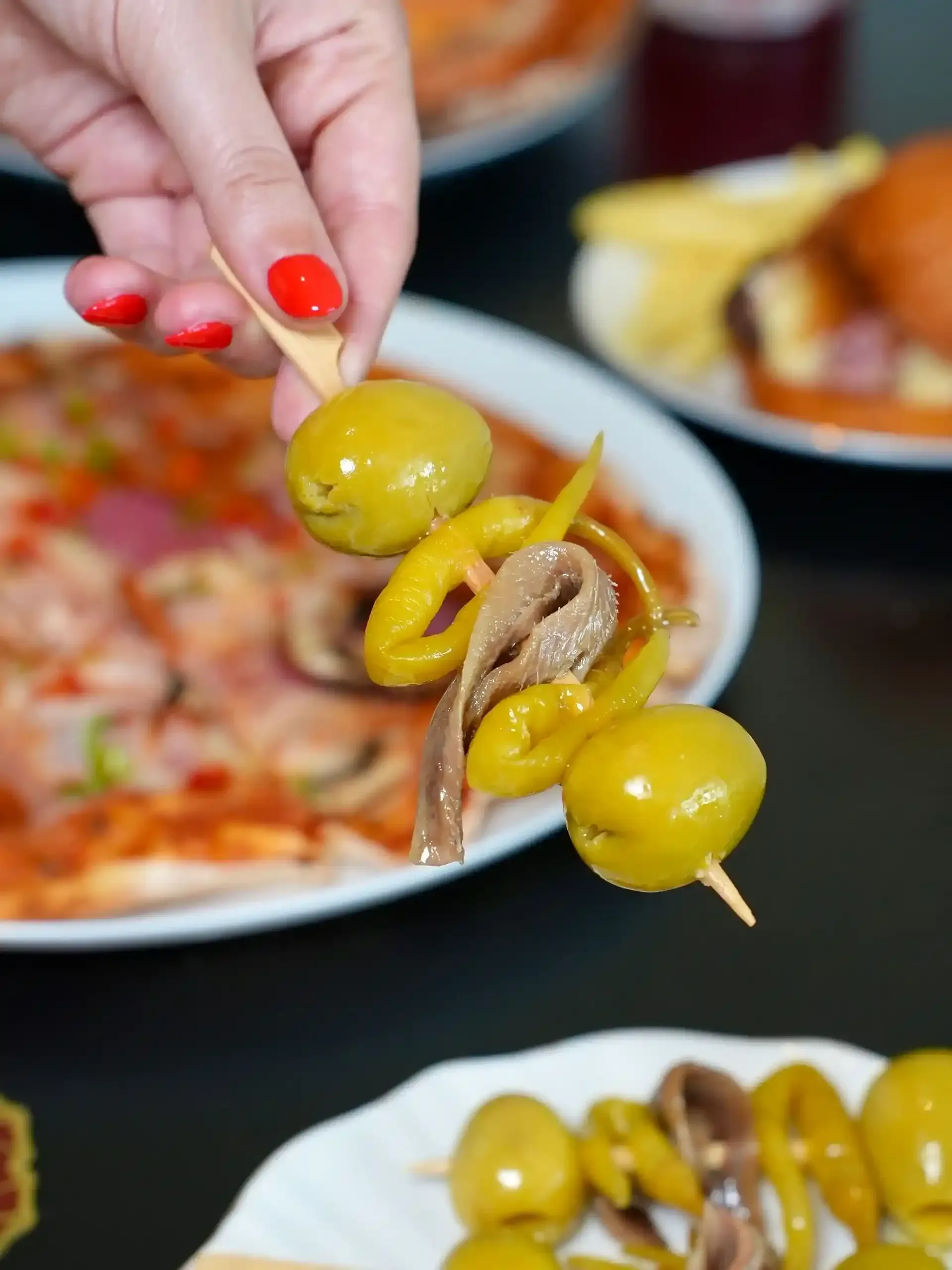 Mano sosteniendo una brocheta con aceitunas verdes, pimientos encurtidos y anchoa. Platos de comida al fondo.