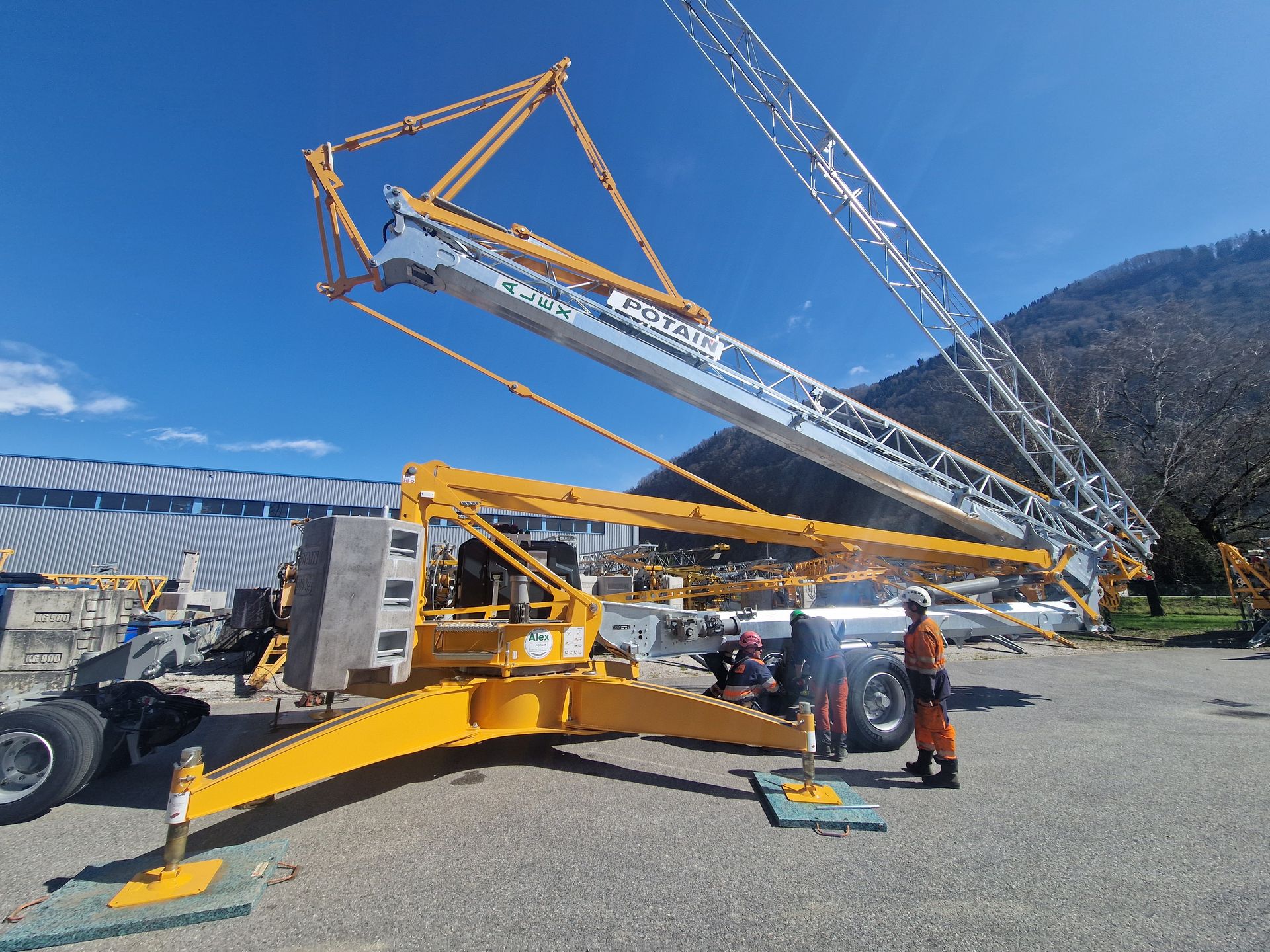 Une grue jaune est déployée, avec un ouvrier à son bord, près d'un bâtiment et de montagnes.