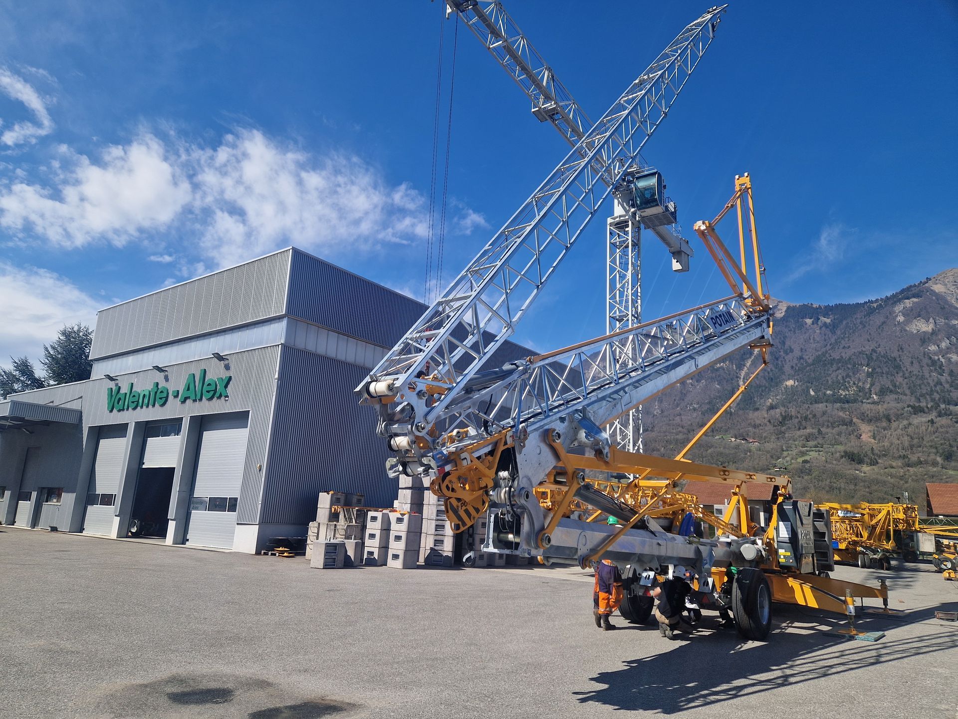 Une grande grue sur un chantier de construction, avec un bâtiment, des ouvriers et des montagnes en arrière-plan.
