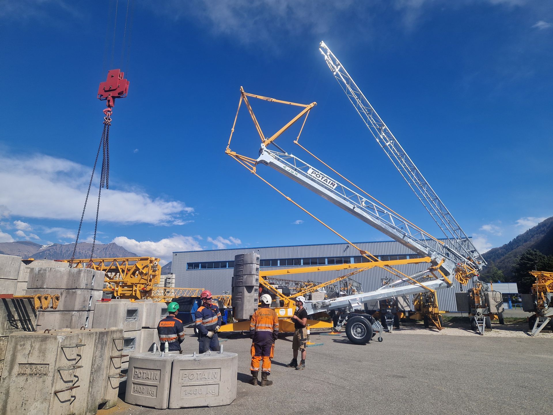 Une grue soulève des blocs de béton près d'une montagne. Des ouvriers en gilet de sécurité observent.