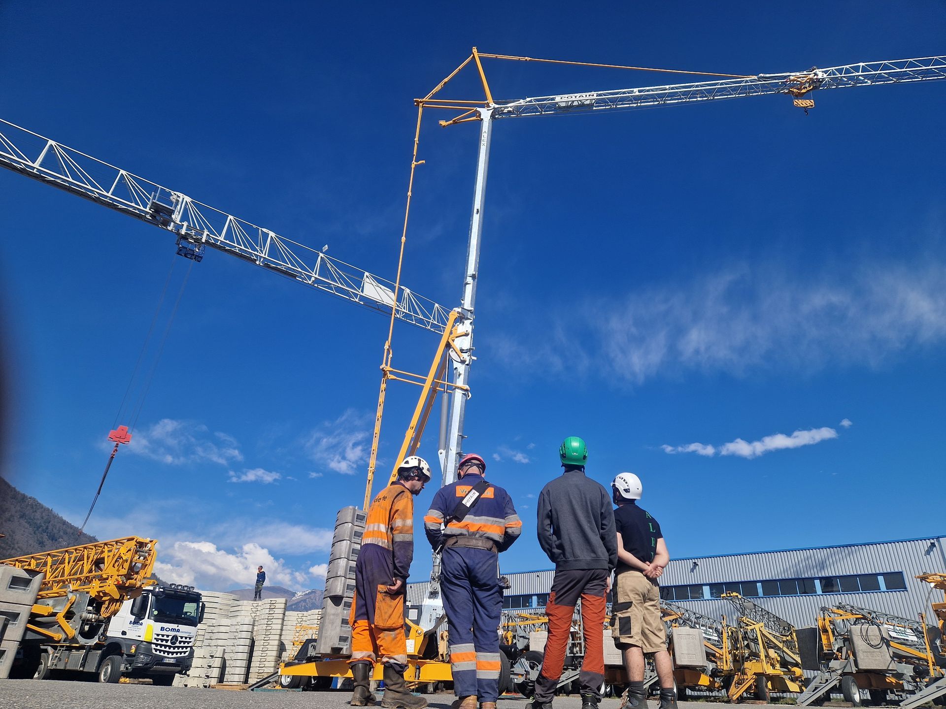 Des ouvriers du bâtiment et des grandes grues sous un ciel bleu. Un camion est stationné à gauche.