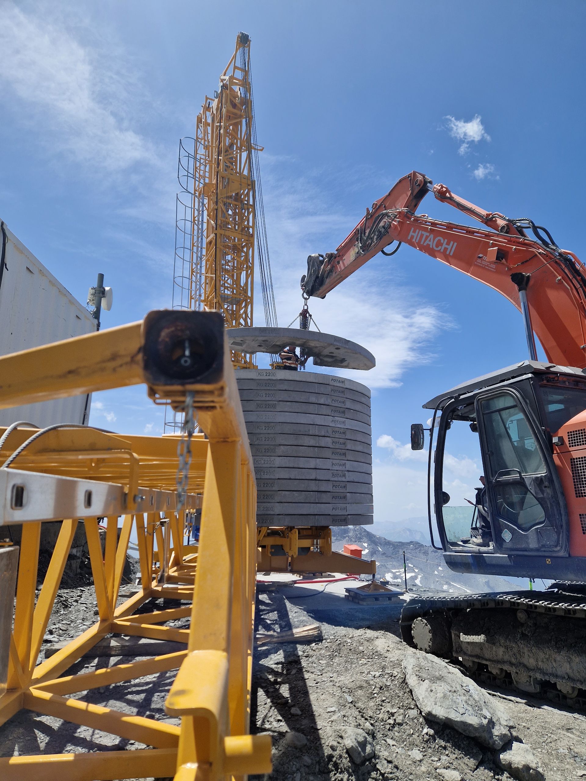 Une excavatrice soulève un anneau de béton, près d'une grande grue de chantier jaune.