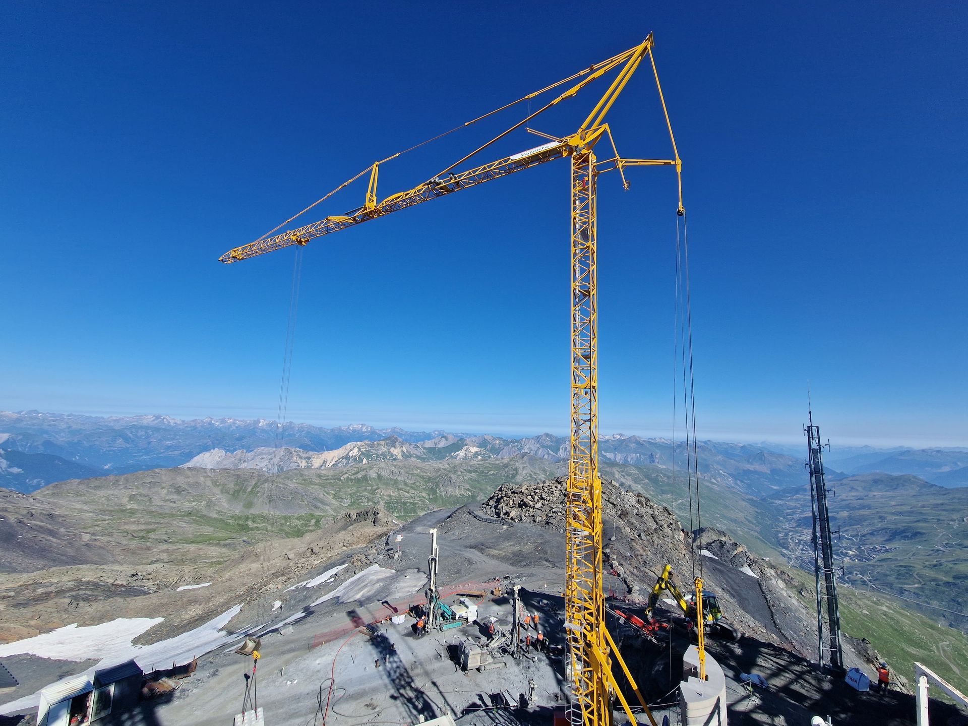 Grue jaune au sommet d'une montagne, ciel bleu en arrière-plan, chantier de construction visible.