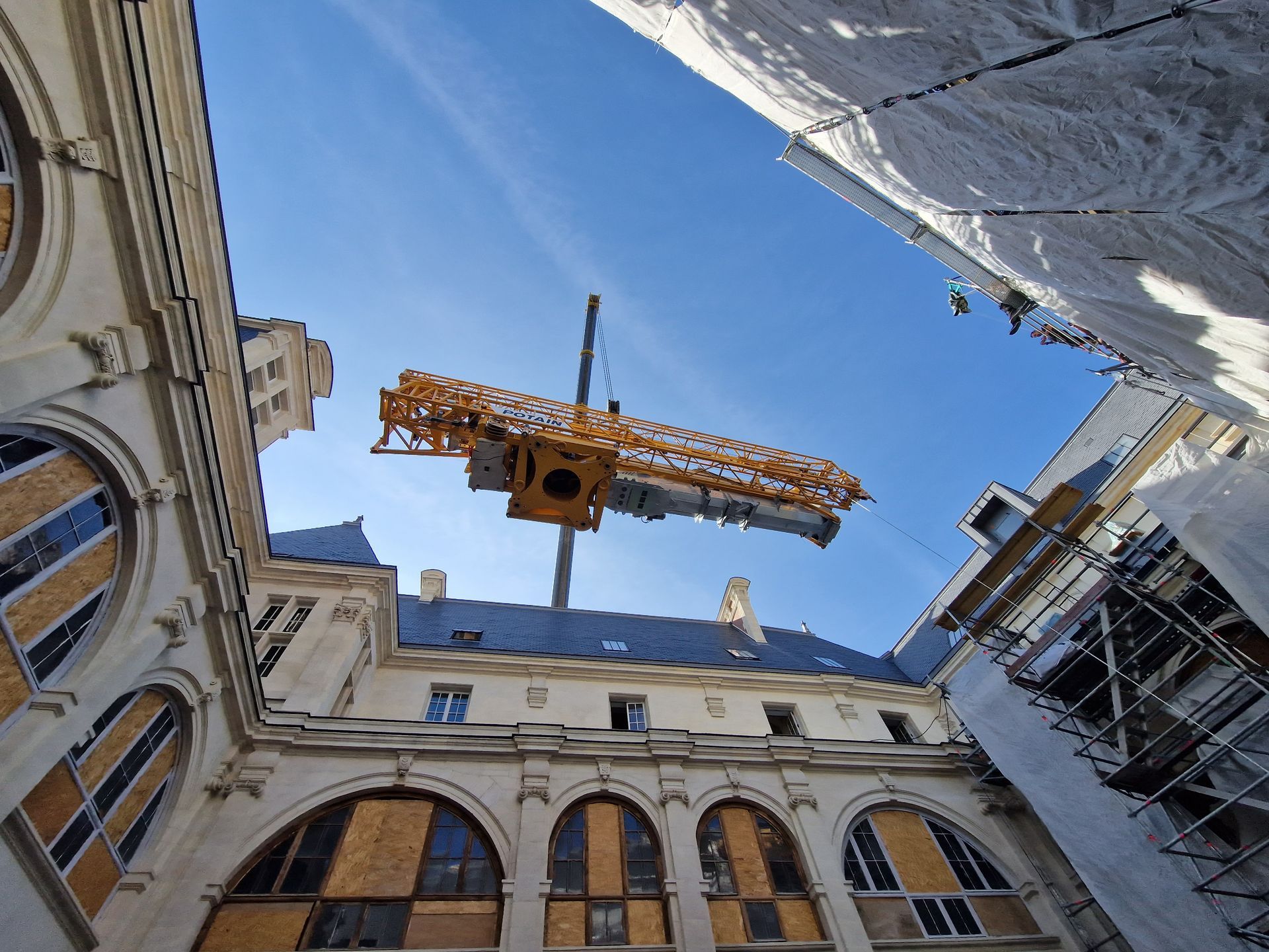 Une grue jaune soulève du matériel au milieu de bâtiments historiques aux fenêtres cintrées, sous un ciel bleu.