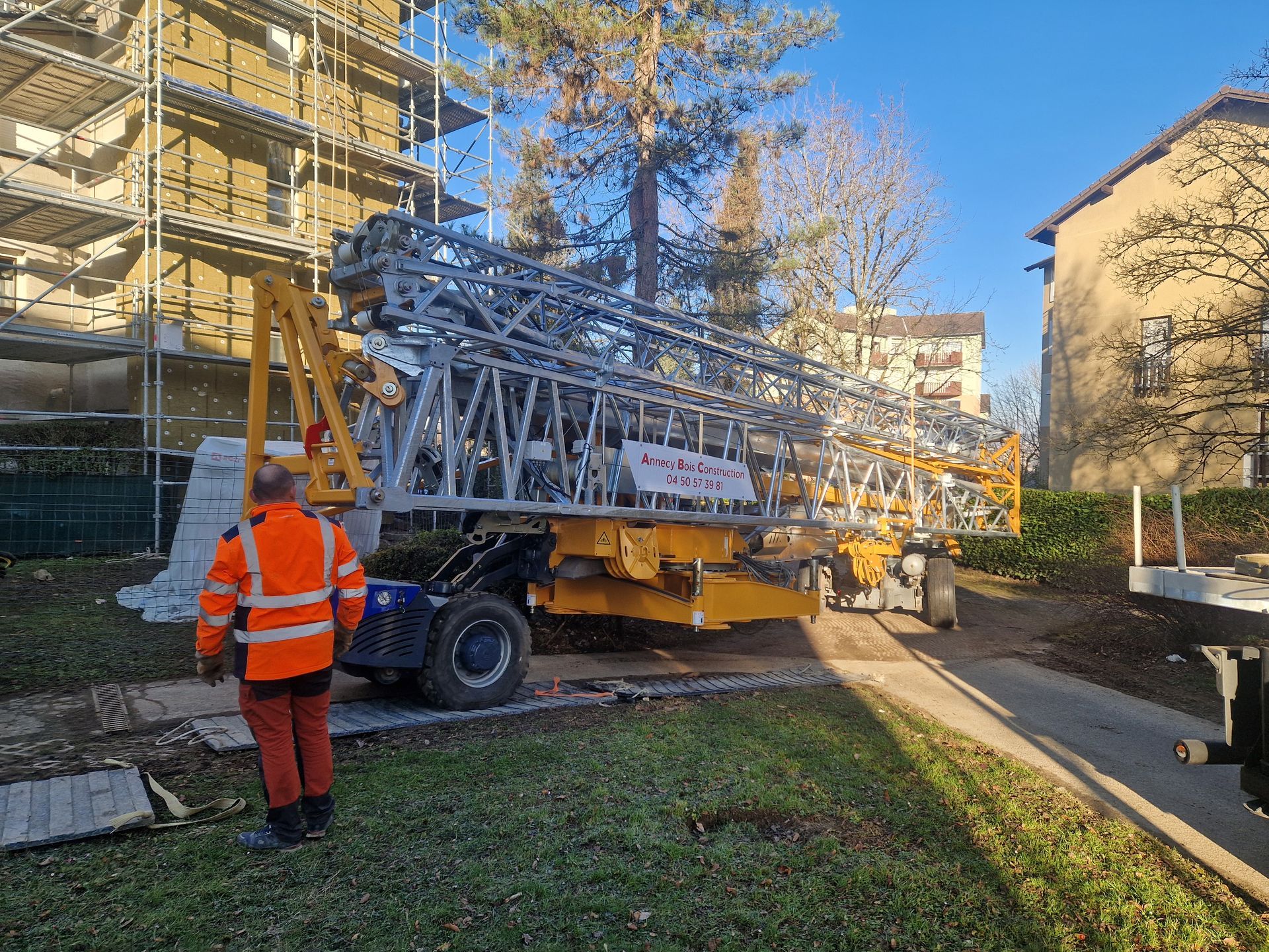 Un ouvrier en gilet orange se trouve près d'un camion transportant une grande grue.