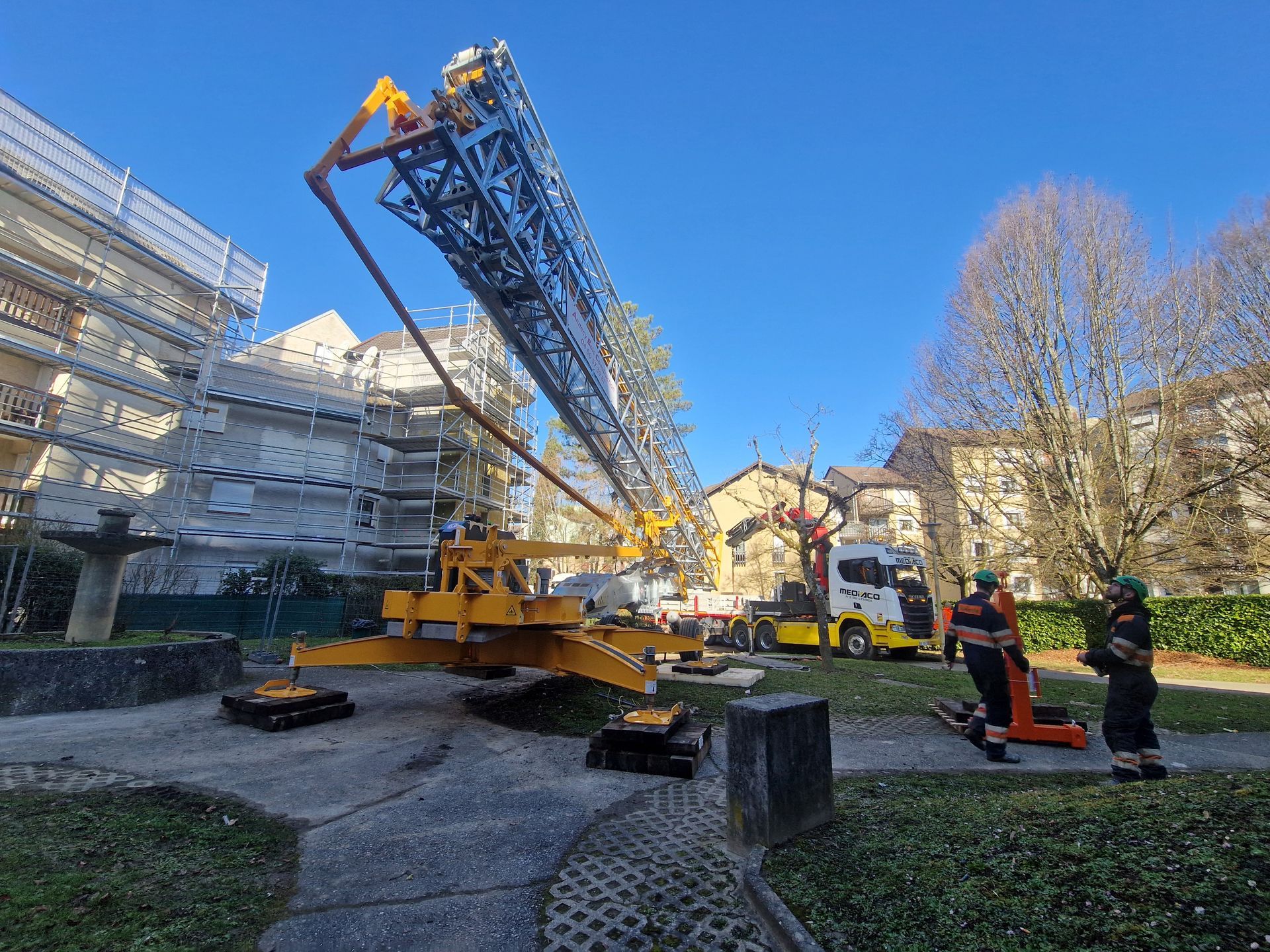Une grande grue jaune s'étend vers un bâtiment. Des ouvriers se tiennent à proximité.