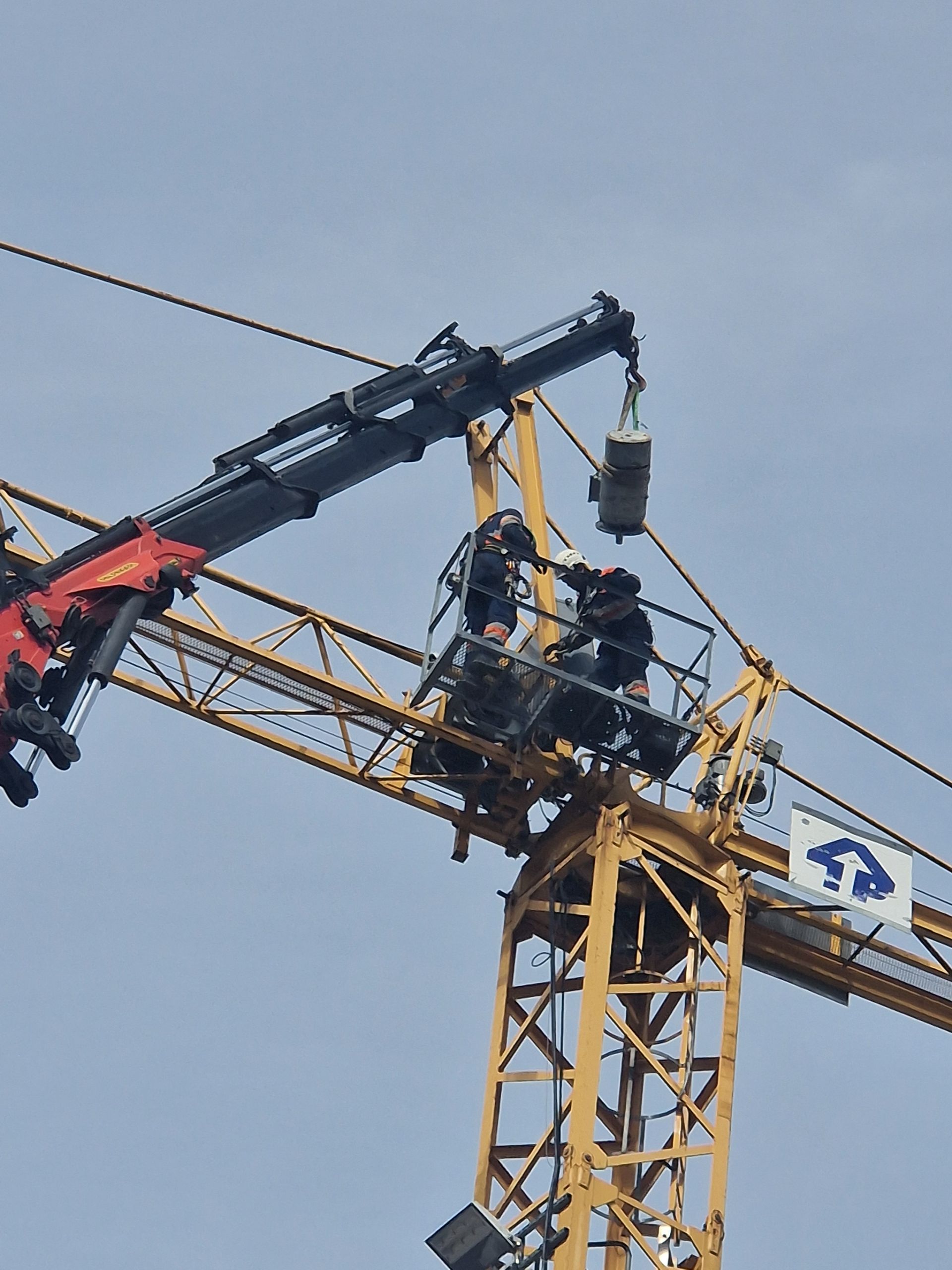 Deux ouvriers sur une grue de chantier jaune ajustent un objet suspendu sur fond de ciel bleu.