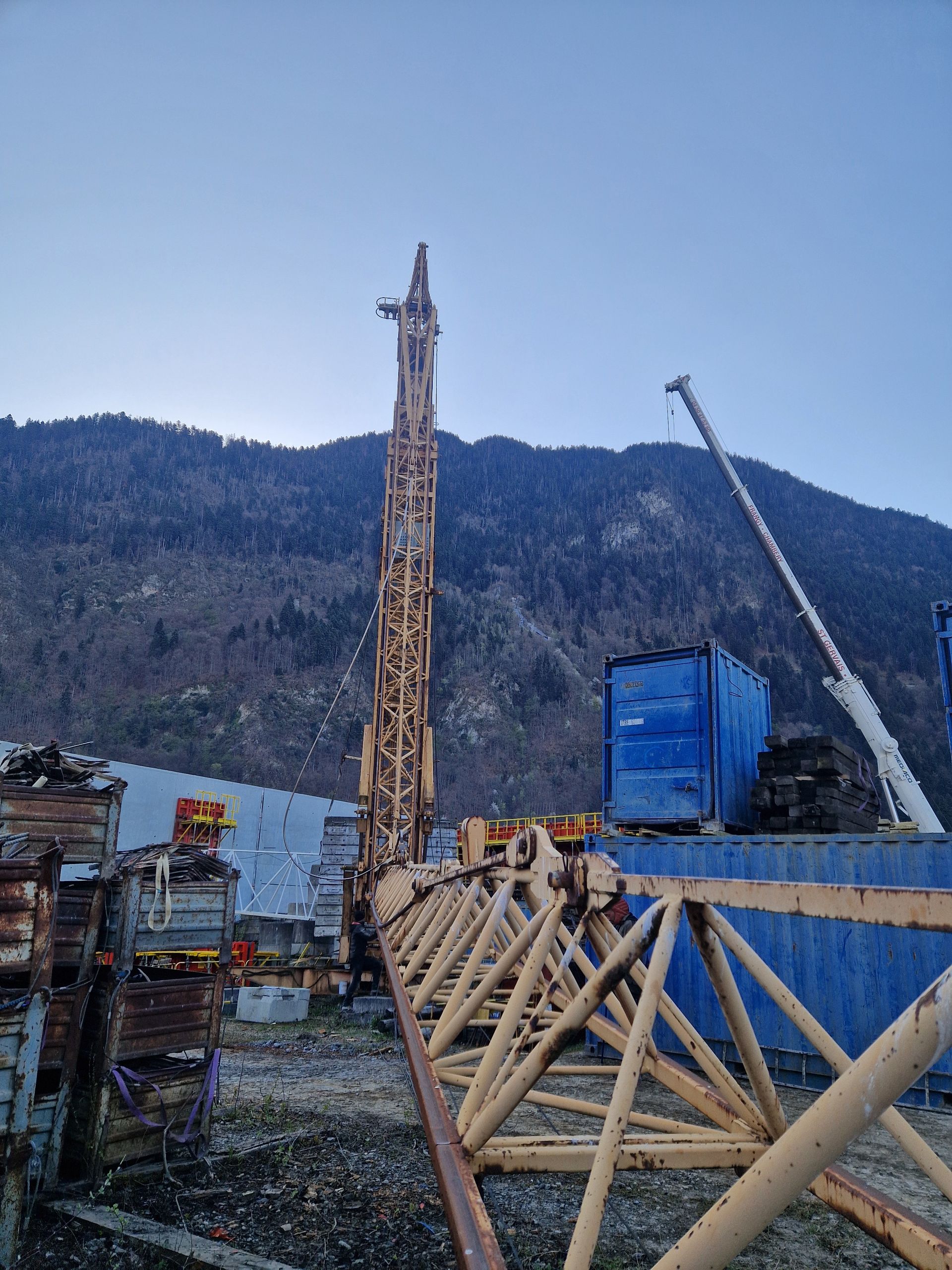 Chantier de construction avec foreuse, grue et conteneur bleu.