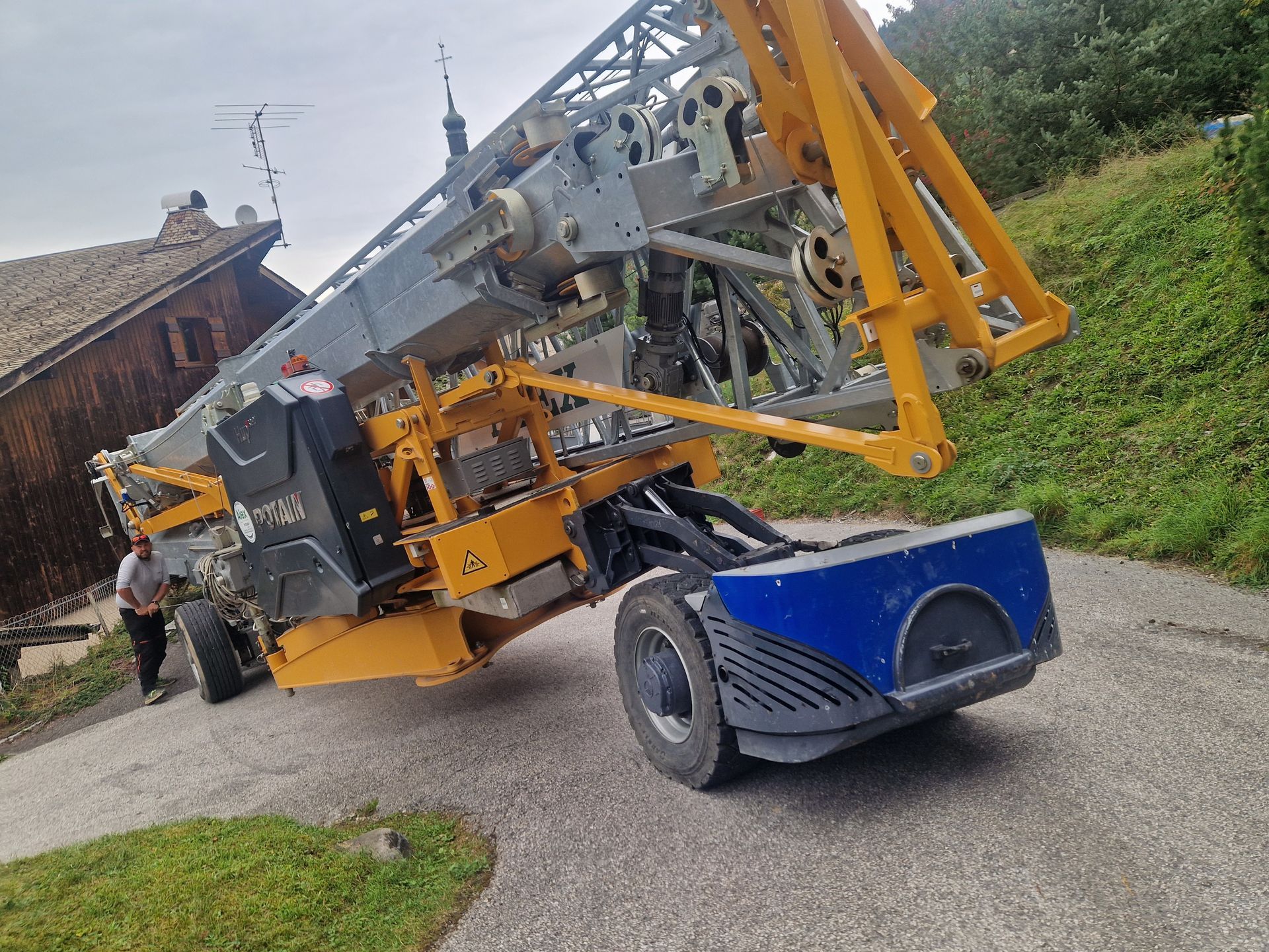 Nacelle élévatrice jaune et bleue sur une route en pente près d'un bâtiment.